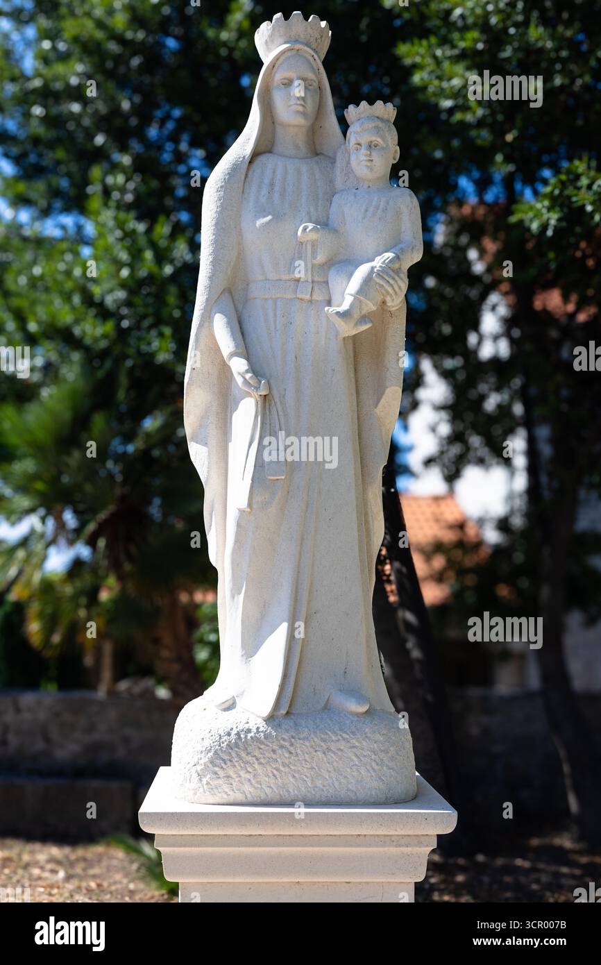 A serene white stone statue of Virgin Mary holding the child, crowned, standing on a pedestal in a sunlit garden. Perfect for religious, memorial, or Stock Photo