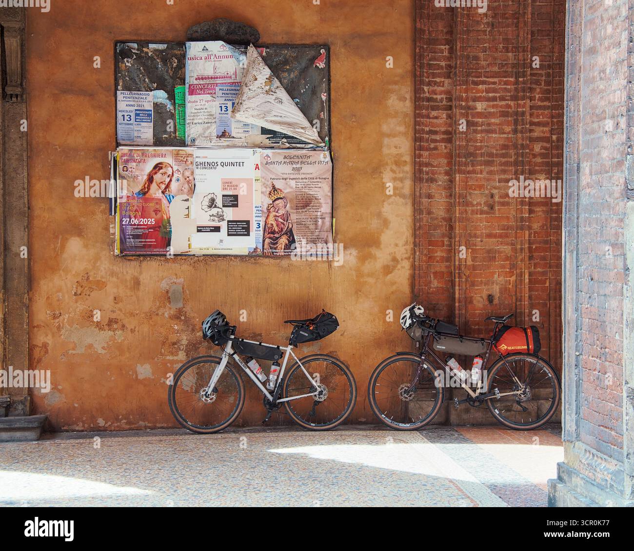 Two bikepacking bicycles under a rustic arcade and notice board, warm ochre wall and brick arch, slow travel in the historic city of Bologna. Stock Photo