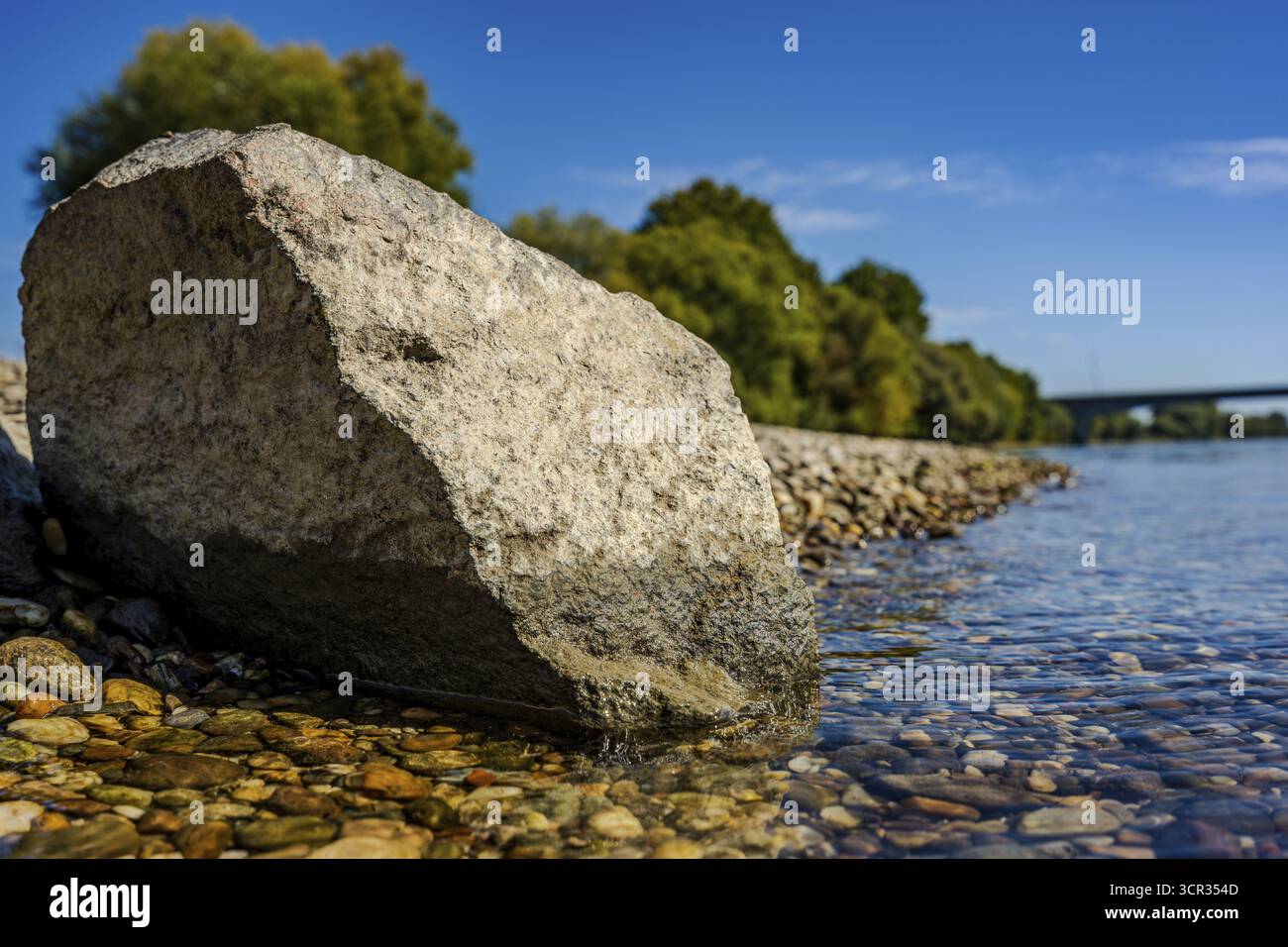 A large stone lies on the bank of a calm river. The water is clear and ...