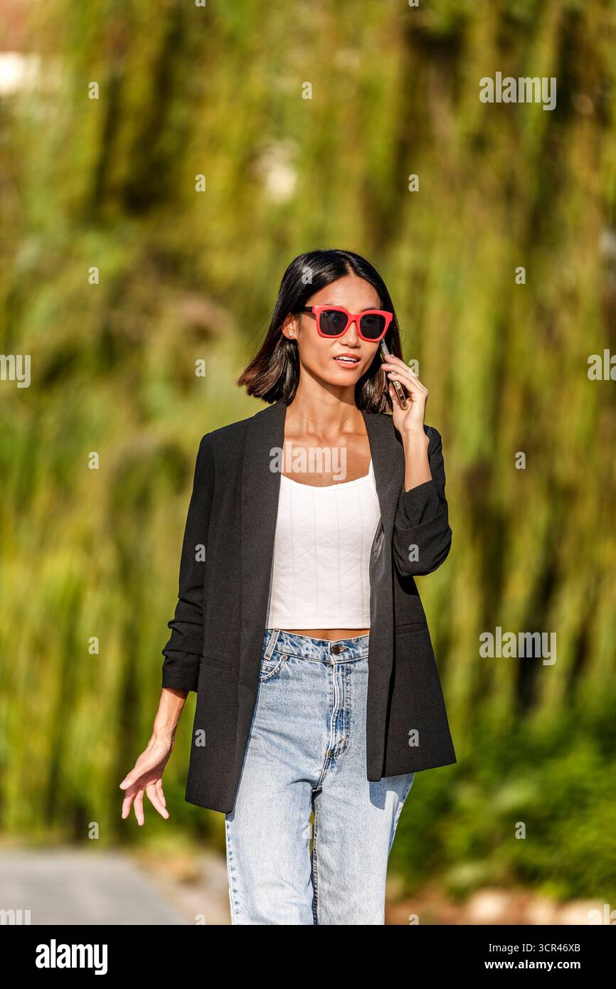 Woman in black blazer and red sunglasses walks while talking on phone outdoors. Spain Stock Photo