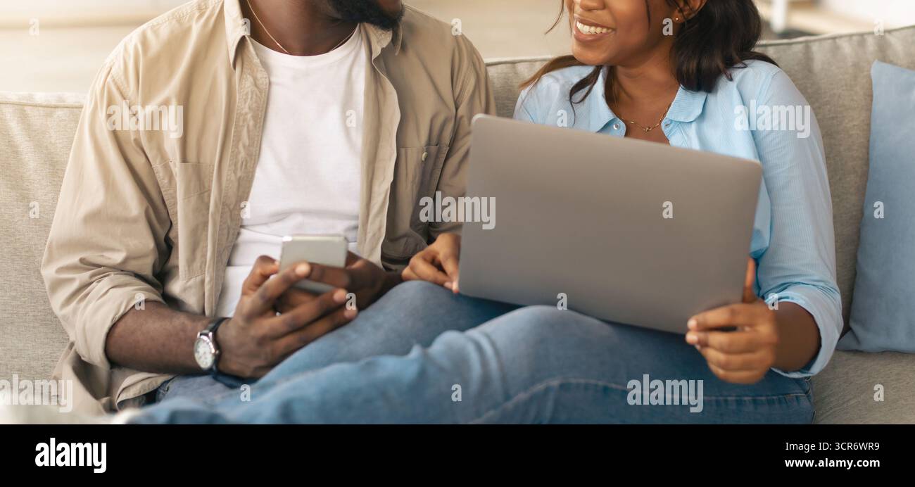 Happy black couple sitting on couch, using cellphone and pc Stock Photo