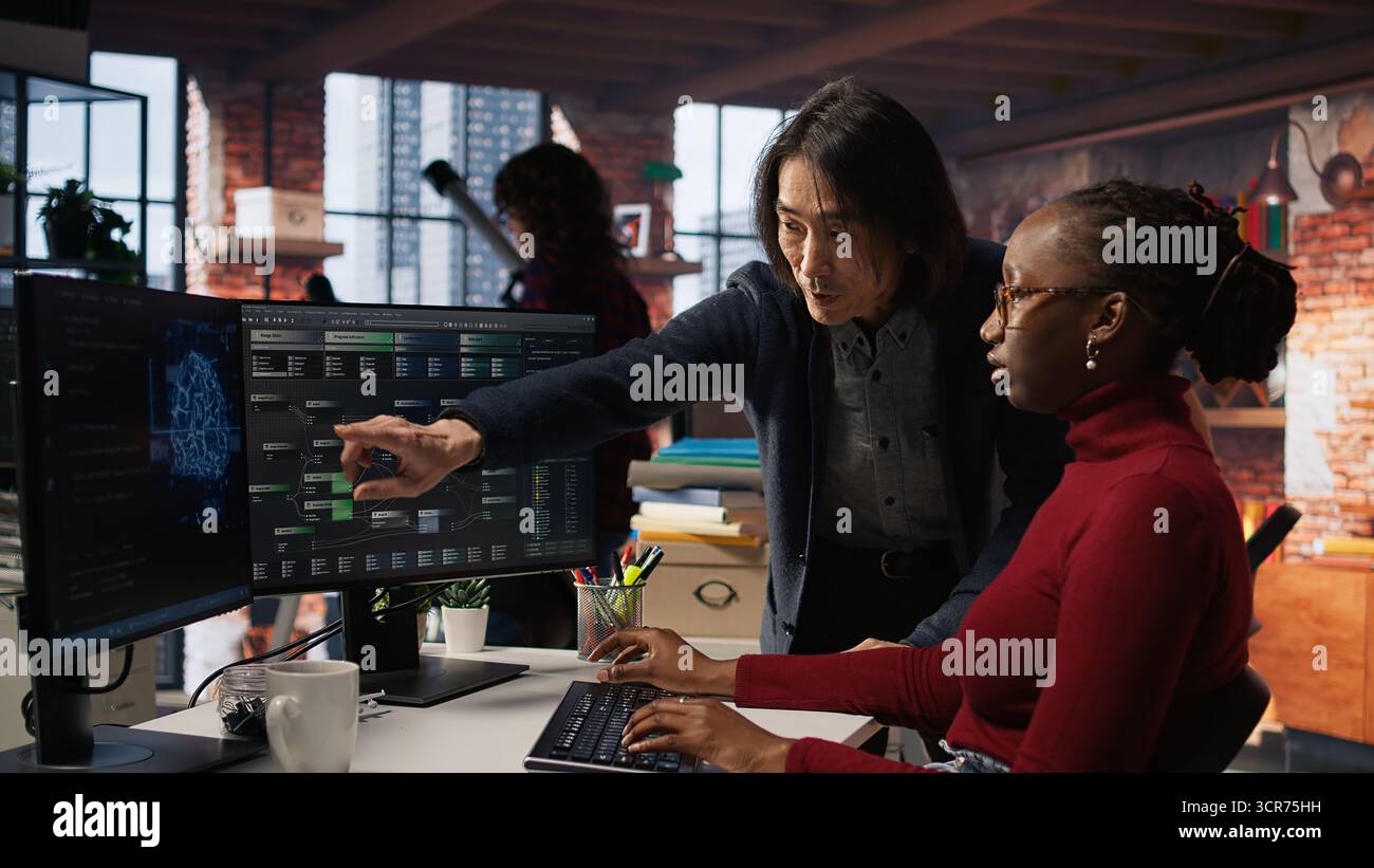 Man in AI startup office CTO position overseeing software engineer implementing machine learning. Artificial intelligence tech venture supervisor helps employee fix code bugs, camera A Stock Photo