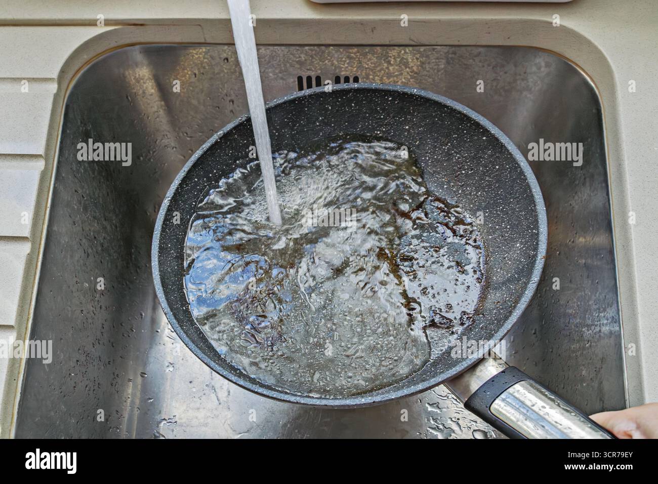 Nonstick pan held under running tap in sink, foam and bits of char washing away, active rinsing Stock Photo
