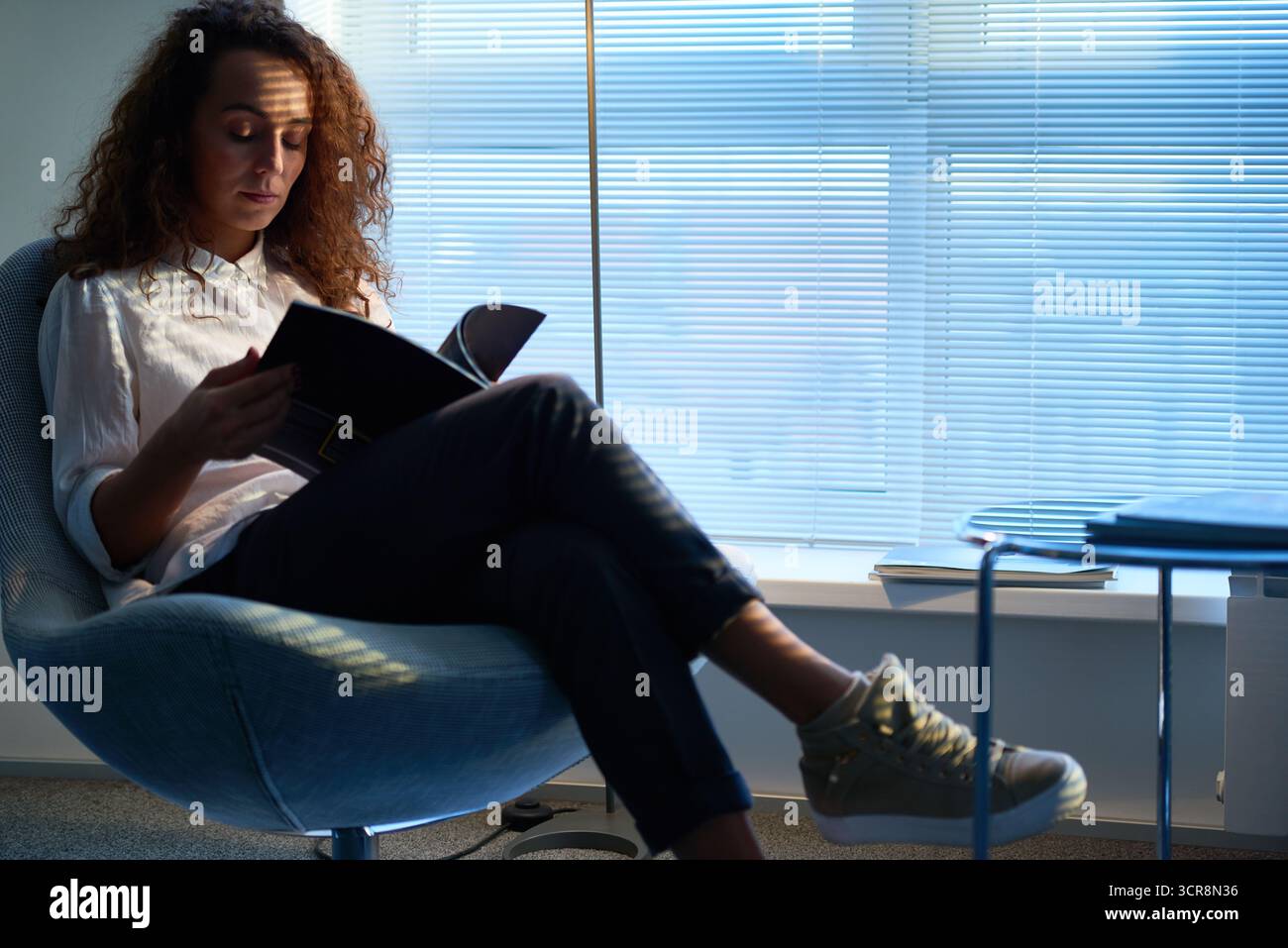 Pretty curly woman wearing white shirt and beige sneakers sitting in waiting room with Venetian blinds on windows, leafing through pages of fashion magazine Stock Photo