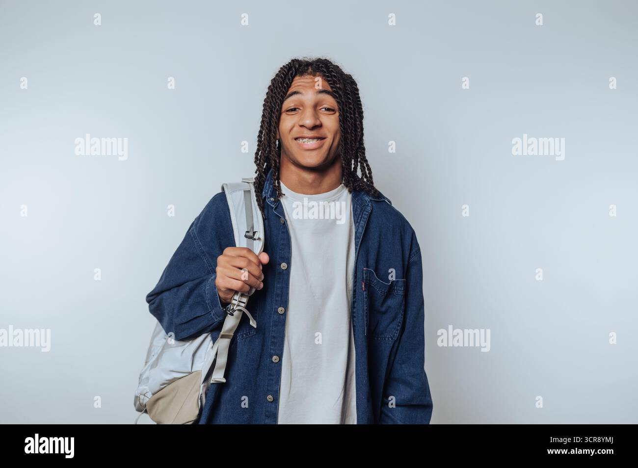 Young man with braided hair smiling and carrying backpack stands against a plain background Stock Photo