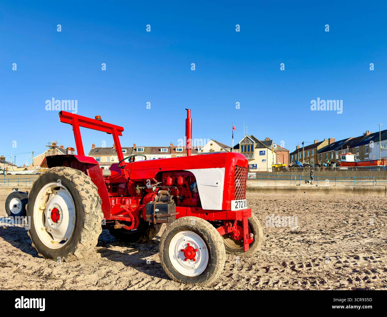 Vintage red David Brown tractor parked on a sandy beach with coastal town buildings and bright blue sky in the background - Smartphone Captured Stock Image