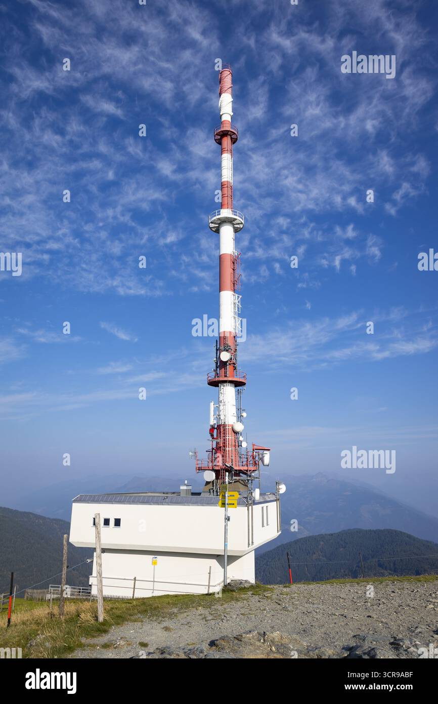 View of a tall, red and white communication tower rises against a clear blue sky dotted with fluffy clouds, Goldeck, Kärnten, Austria. Stock Photo