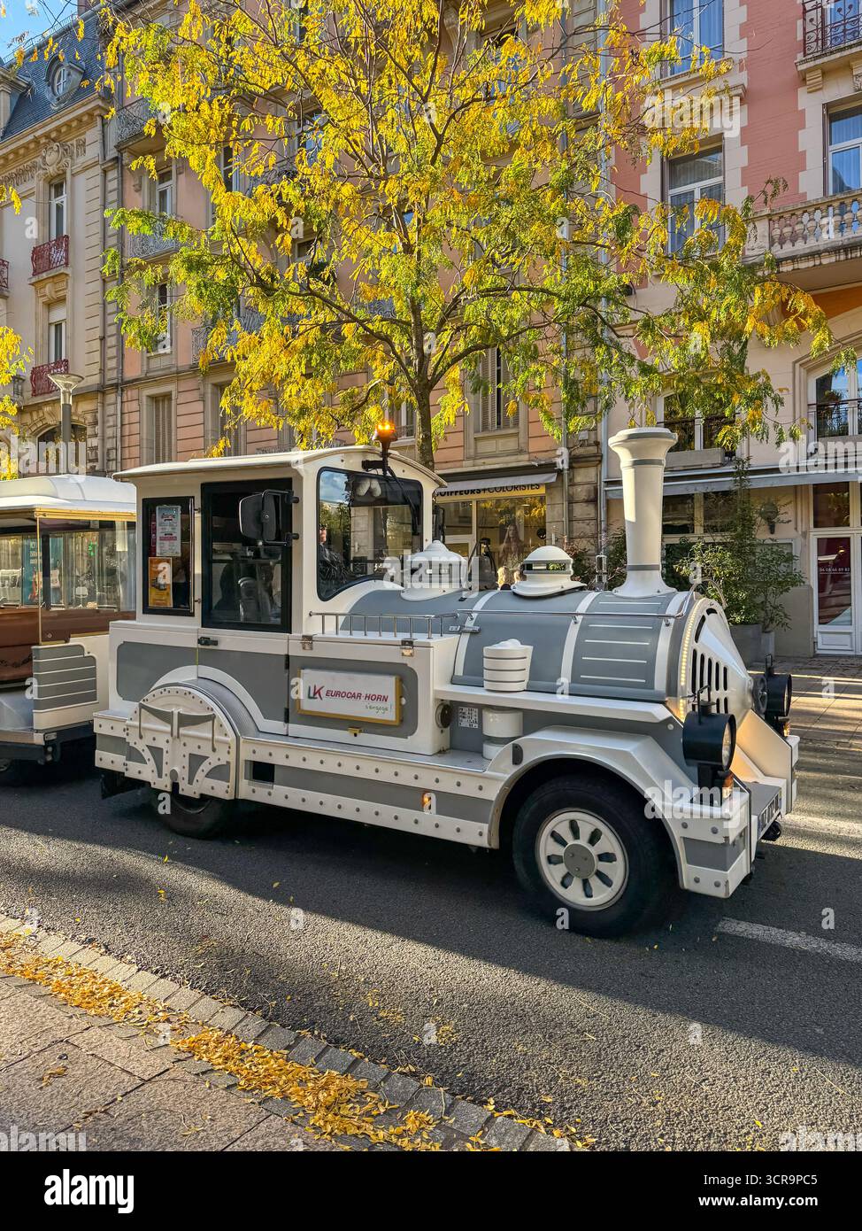 Tourist road  train parked along a tree lined street with autumn foliage, in front of historic european buildings. - Smartphone Captured Stock Image