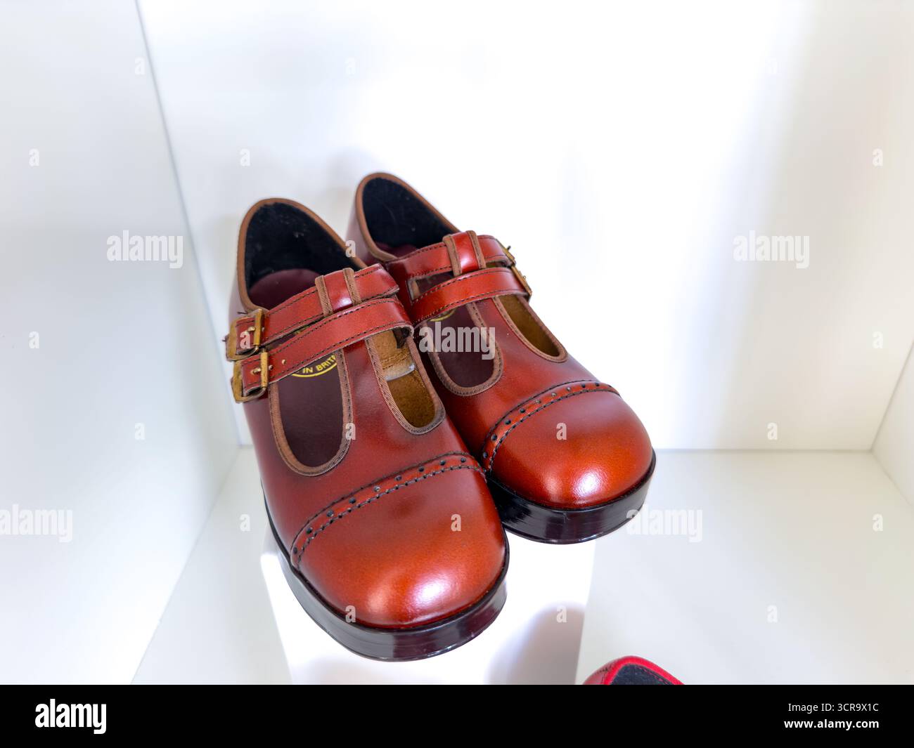 A pair of shiny brown leather Mary Jane shoes with double straps displayed on a white shelf under bright lighting - Smartphone Captured Stock Image
