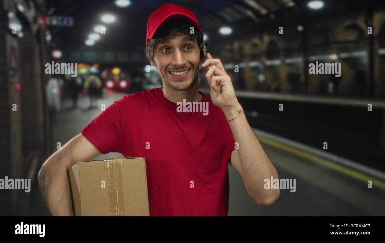 Man holds cardboard box with one arm and talks on phone at train station; efficiency reliability service. Stock Photo