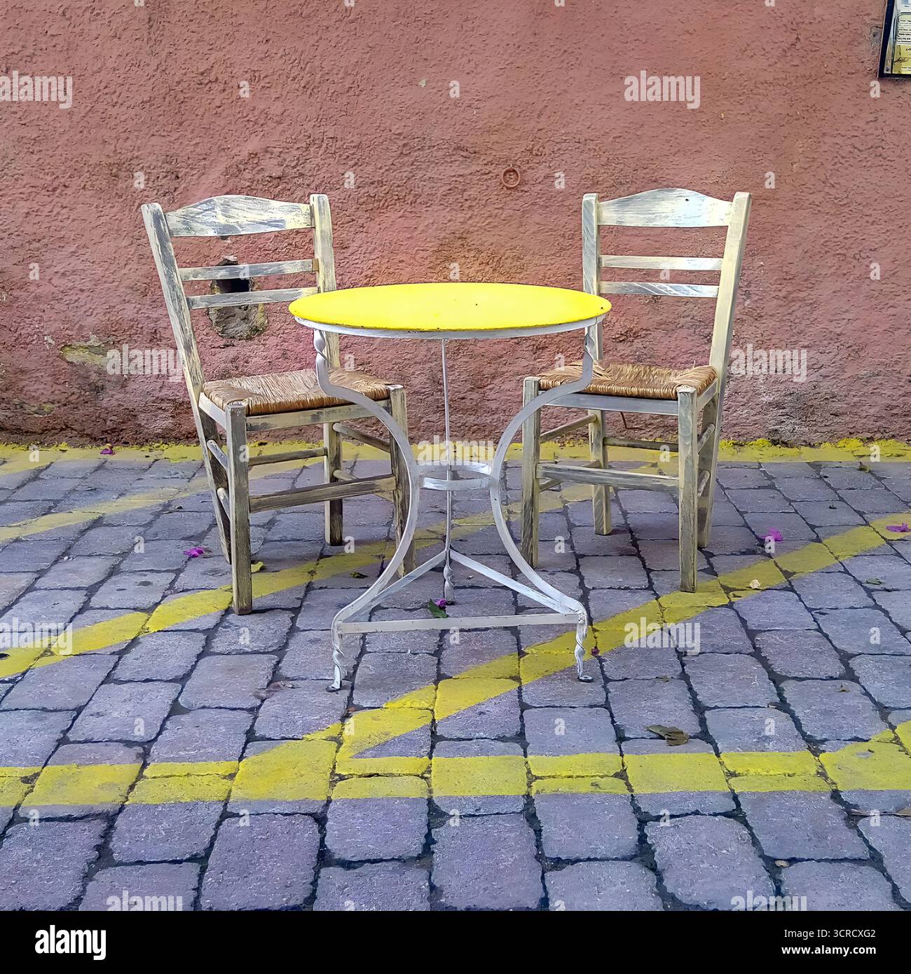 Rustic café set with two weathered chairs and small yellow round table on cobblestone street. Stock Photo