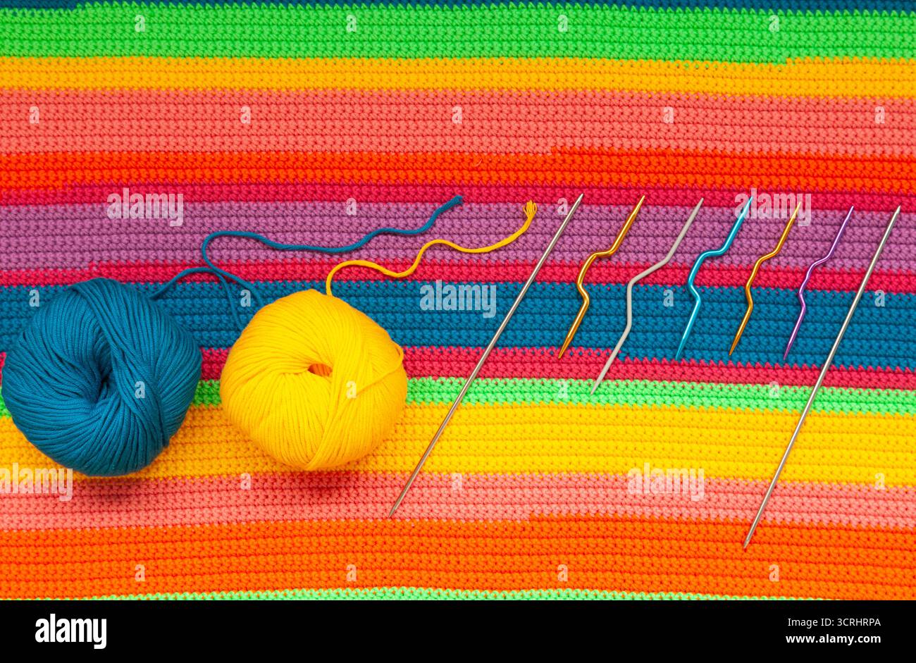 Knitted multi-colored handmade fabric with balls of yellow, blue threads and knitting needles. Stock Photo