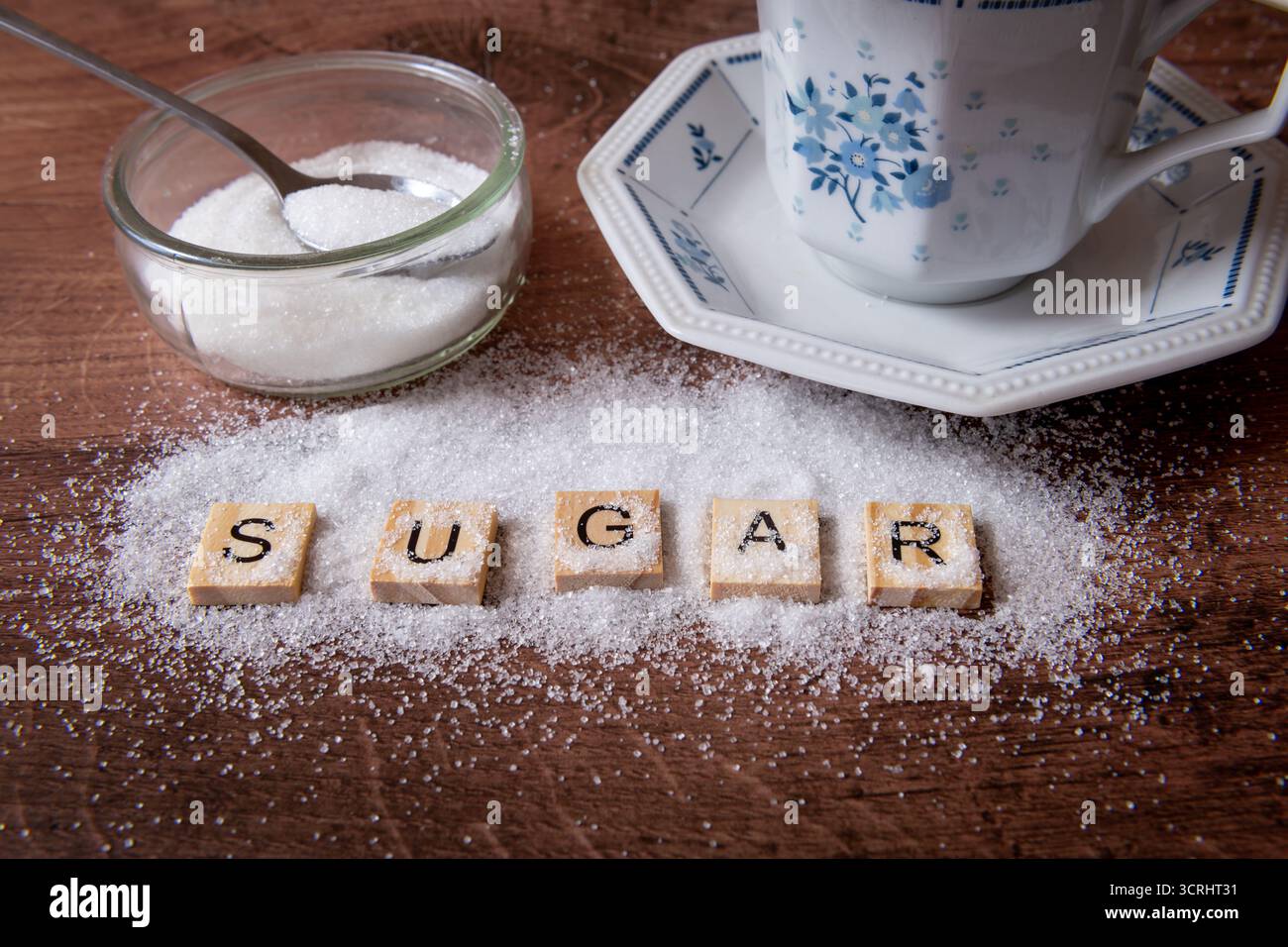 The word sugar with a sprinkle of white sugar together with a tea cup and saucer, and sugar and tea spoon in a glass container. Stock Photo