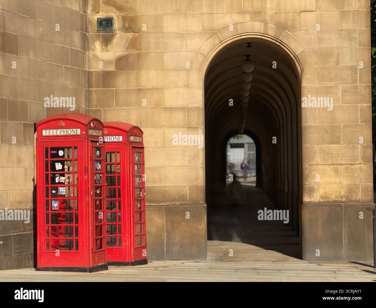 Red traditional telephone kiosks in front of arches of passage in  Manchester town hall in St Peter’s Square with blurred figures - Smartphone Captured Stock Image