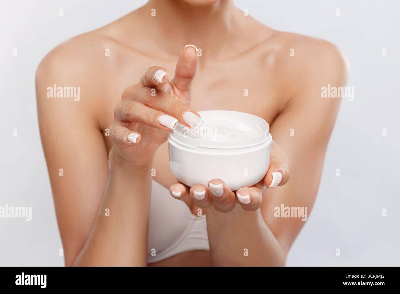 A woman with well-groomed nails is using a finger to scoop moisturizer from a jar. She appears focused and is in a well-lit environment, possibly in a Stock Photo