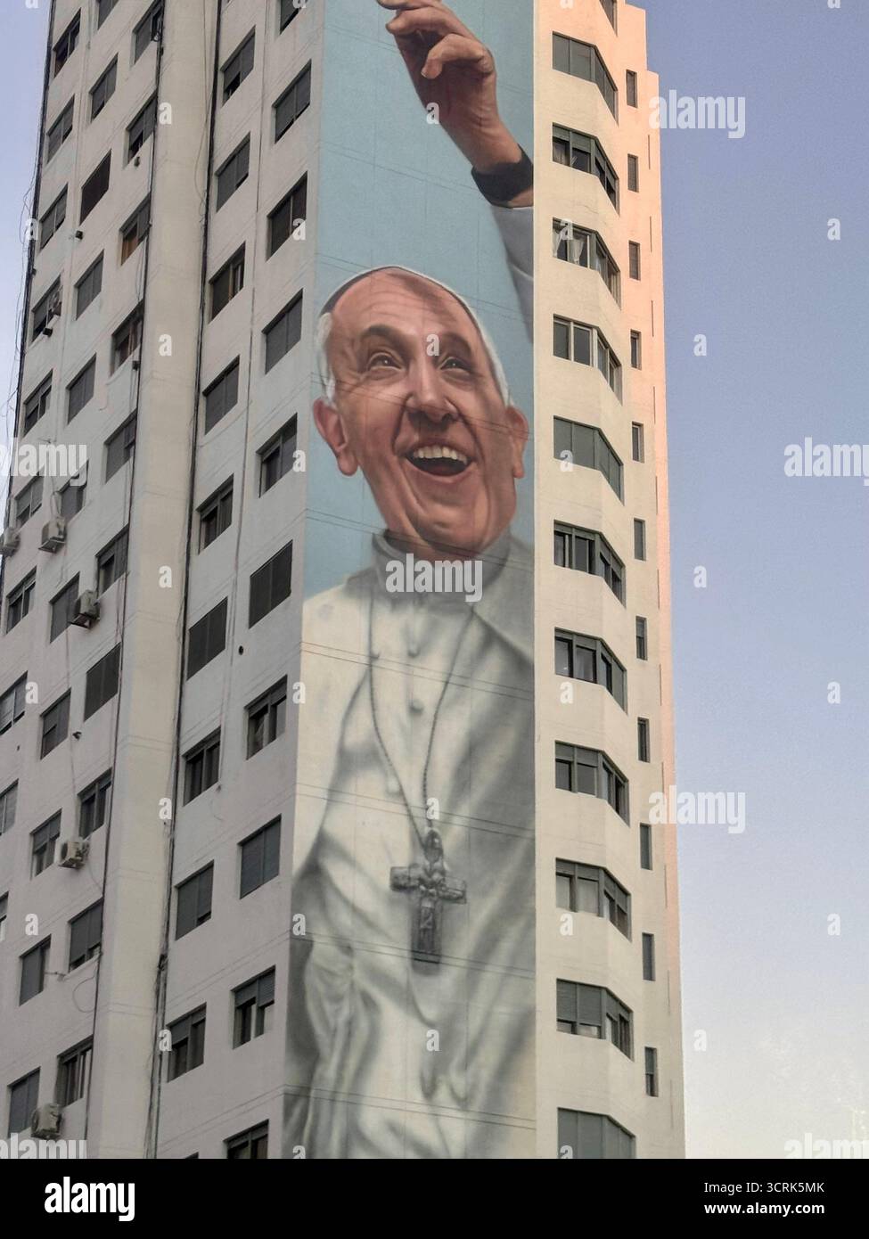 A tall apartment building features a large mural of Pope Francis raising his hand painted on its facade, set against a clear blue sky. - Smartphone Captured Stock Image