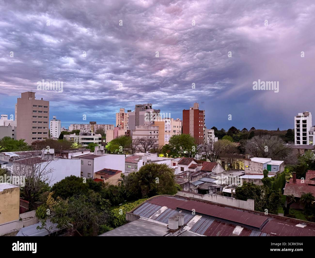 An urban cityscape featuring mid-rise apartment buildings and houses surrounded by trees under a dramatic, colorful cloudy sky at sunset. - Smartphone Captured Stock Image