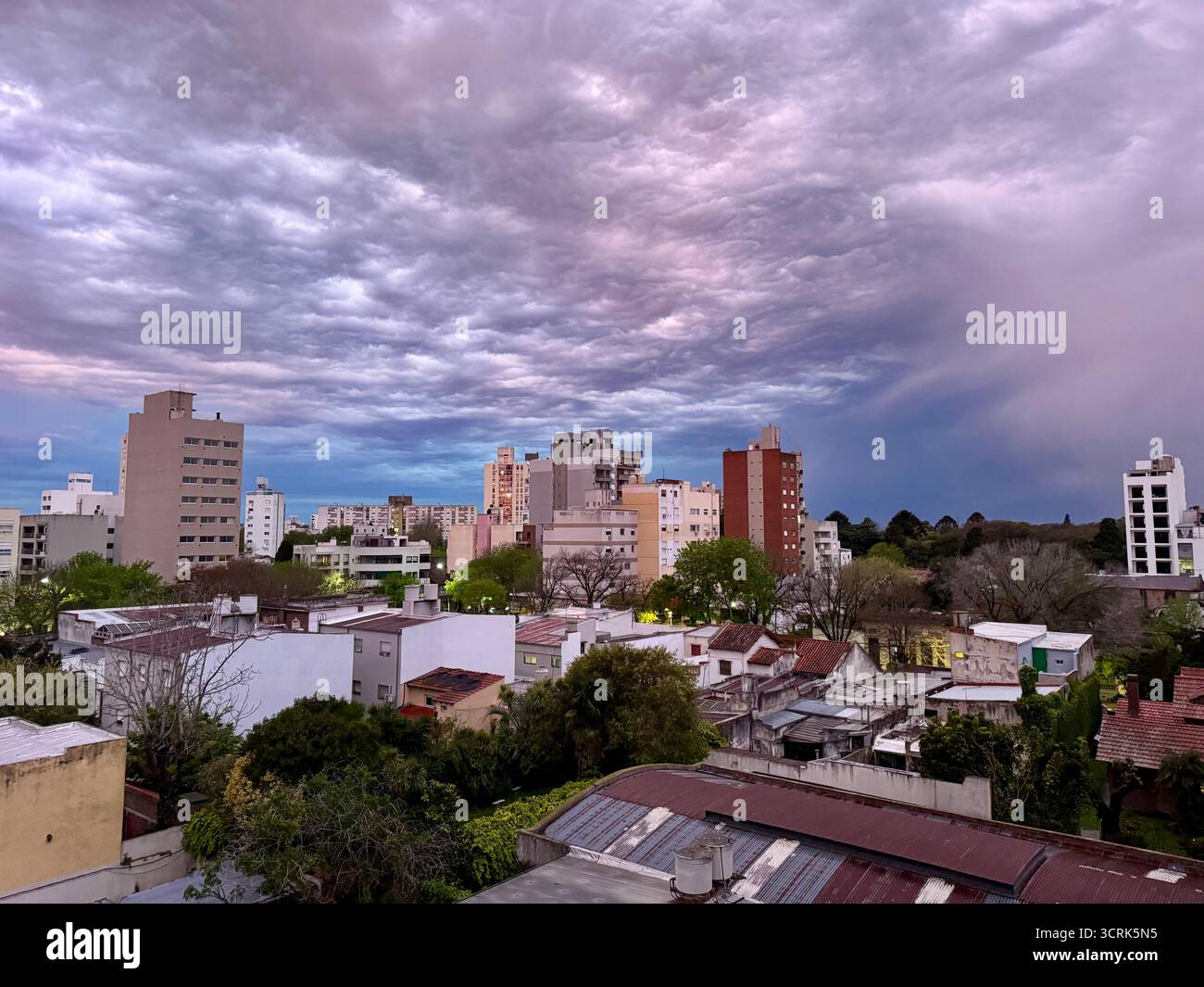 An urban cityscape featuring mid-rise apartment buildings and houses surrounded by trees under a dramatic, colorful cloudy sky at sunset. - Smartphone Captured Stock Image