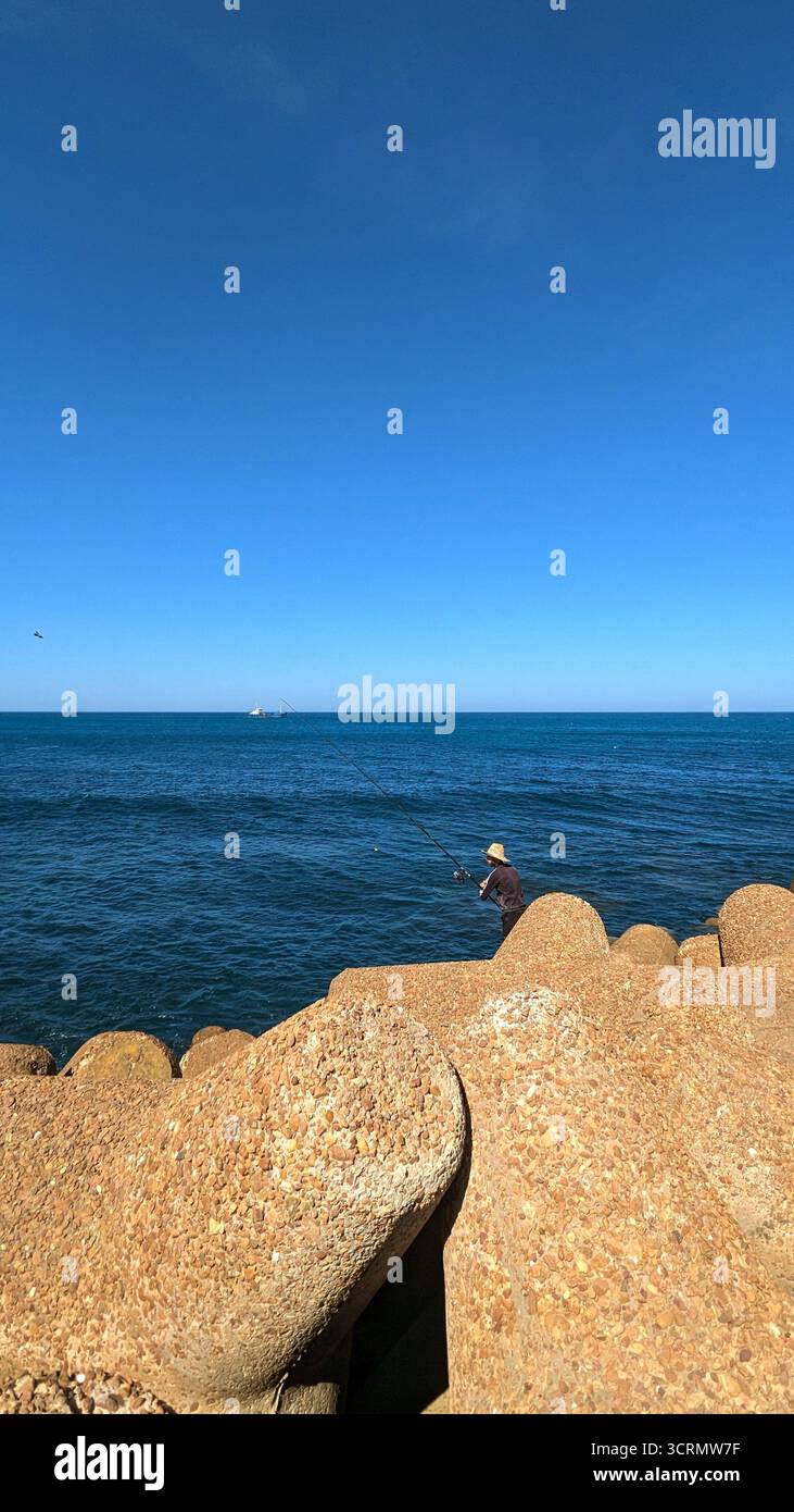 lone fisherman standing on a large concrete breakwaters, casting his line into the deep blue ocean under a clear sunny sky. - Smartphone Captured Stock Image