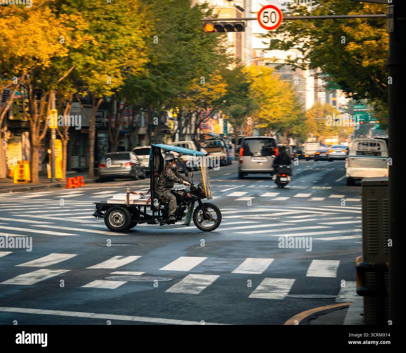 three-wheeled delivery motorcycle carries paper pallets through Chungmuro, Seoul, in golden hour light, casting long shadow across the sunlit asphalte Stock Photo