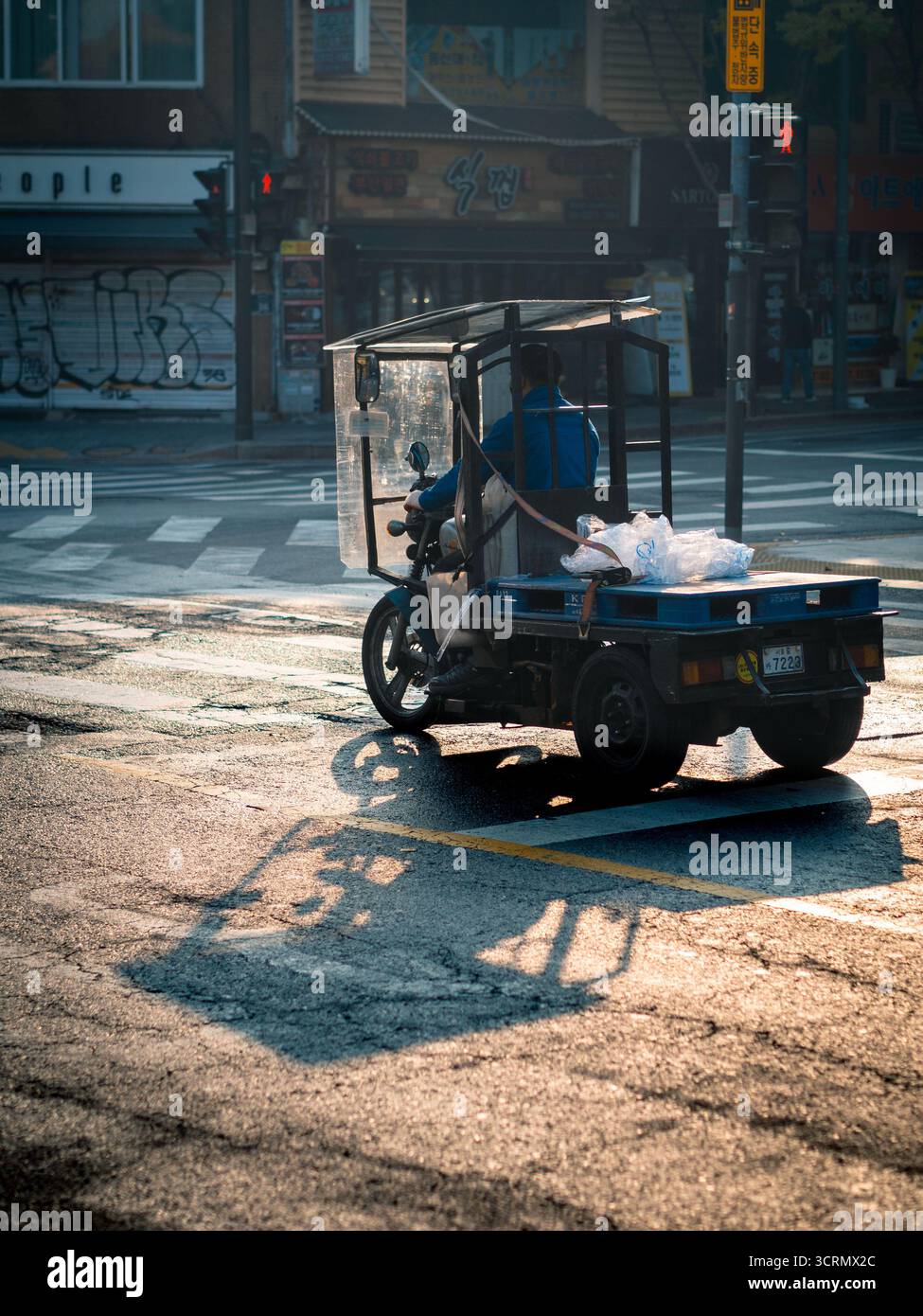 three-wheeled delivery motorcycle carries paper pallets through Chungmuro, Seoul, in golden hour light, casting long shadow across the sunlit asphalte Stock Photo