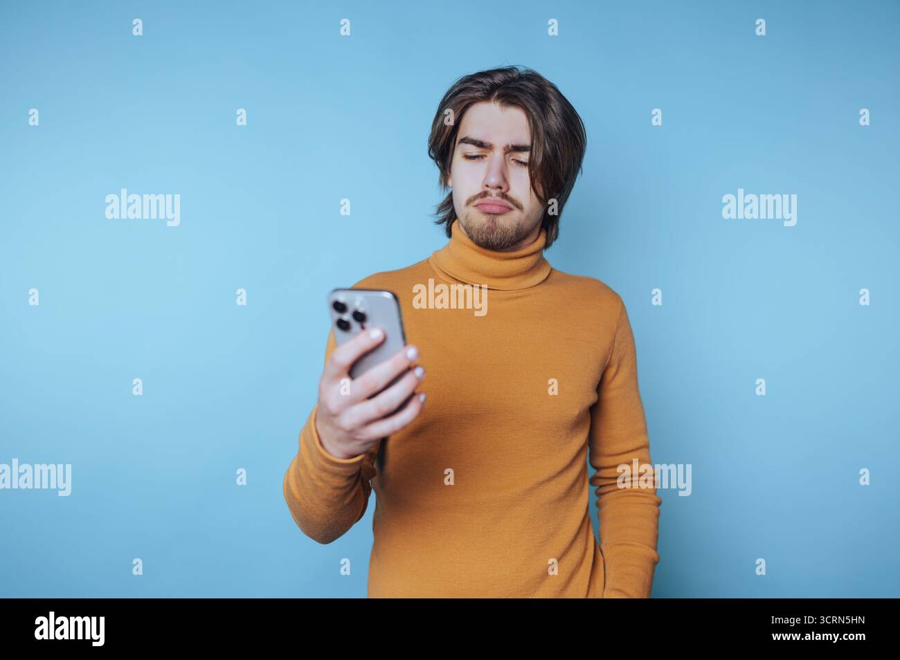 Young man in orange sweater looking at smartphone with puzzled expression. Stock Photo