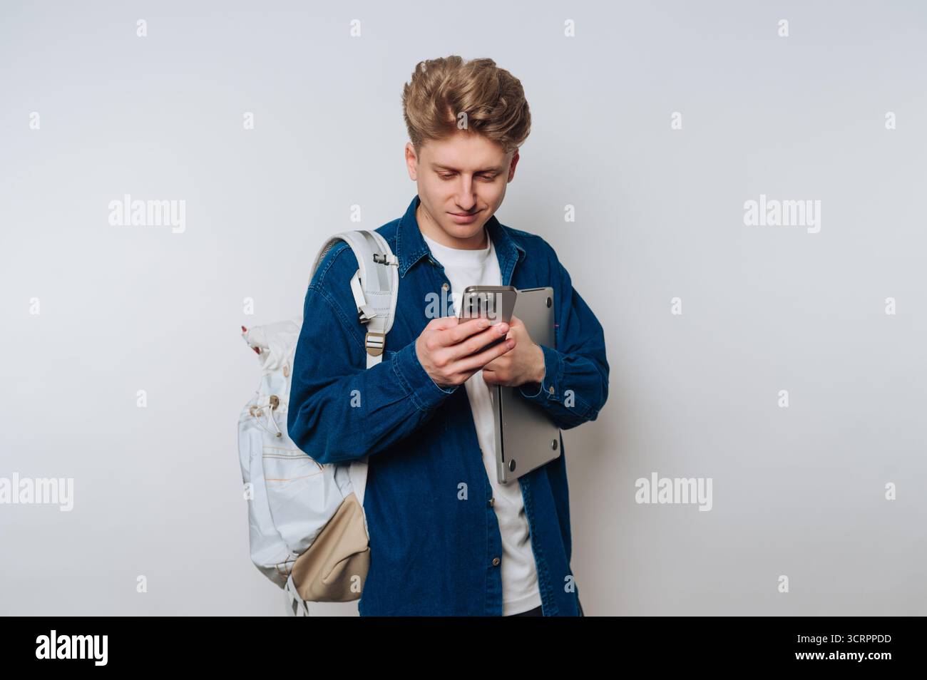 Young man with backpack using smartphone, holding laptop. Stock Photo