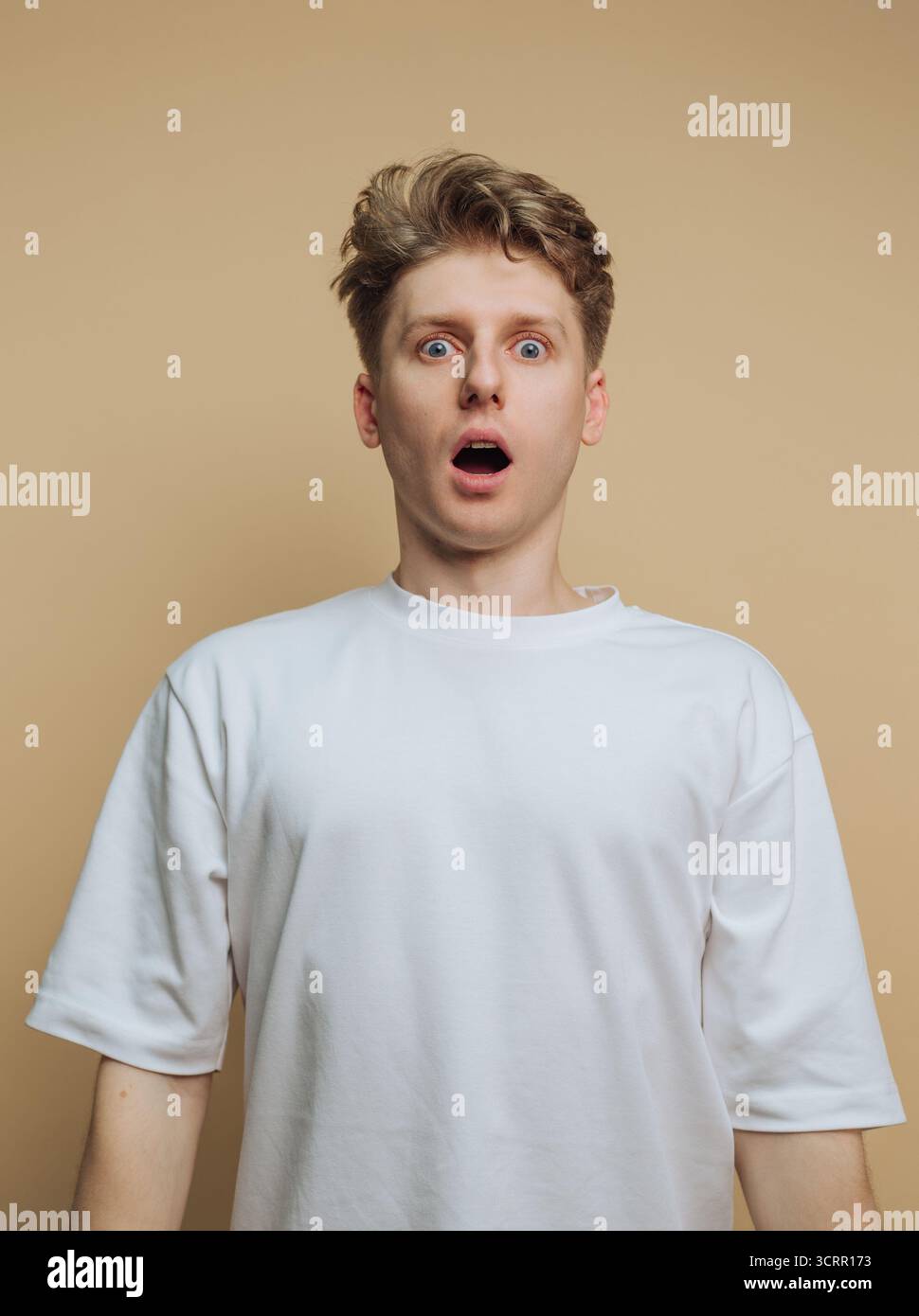 A young man with blonde hair appears surprised, his eyes wide open and mouth agape. He stands against a neutral beige backdrop, wearing a simple white Stock Photo