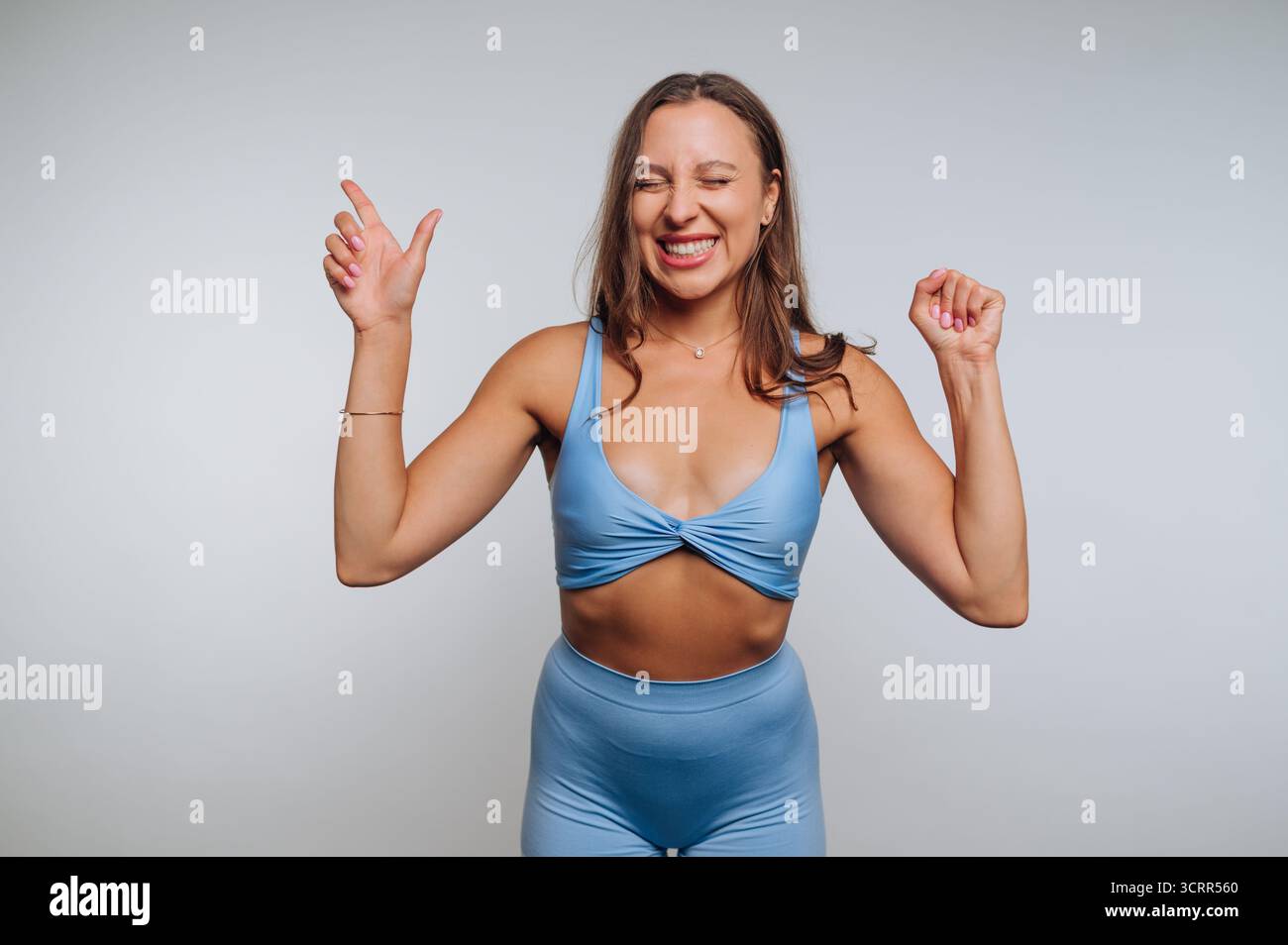 A young woman wearing blue workout clothes joyfully raises her hands and smiles. She appears energized and happy, capturing the moment in a bright stu Stock Photo
