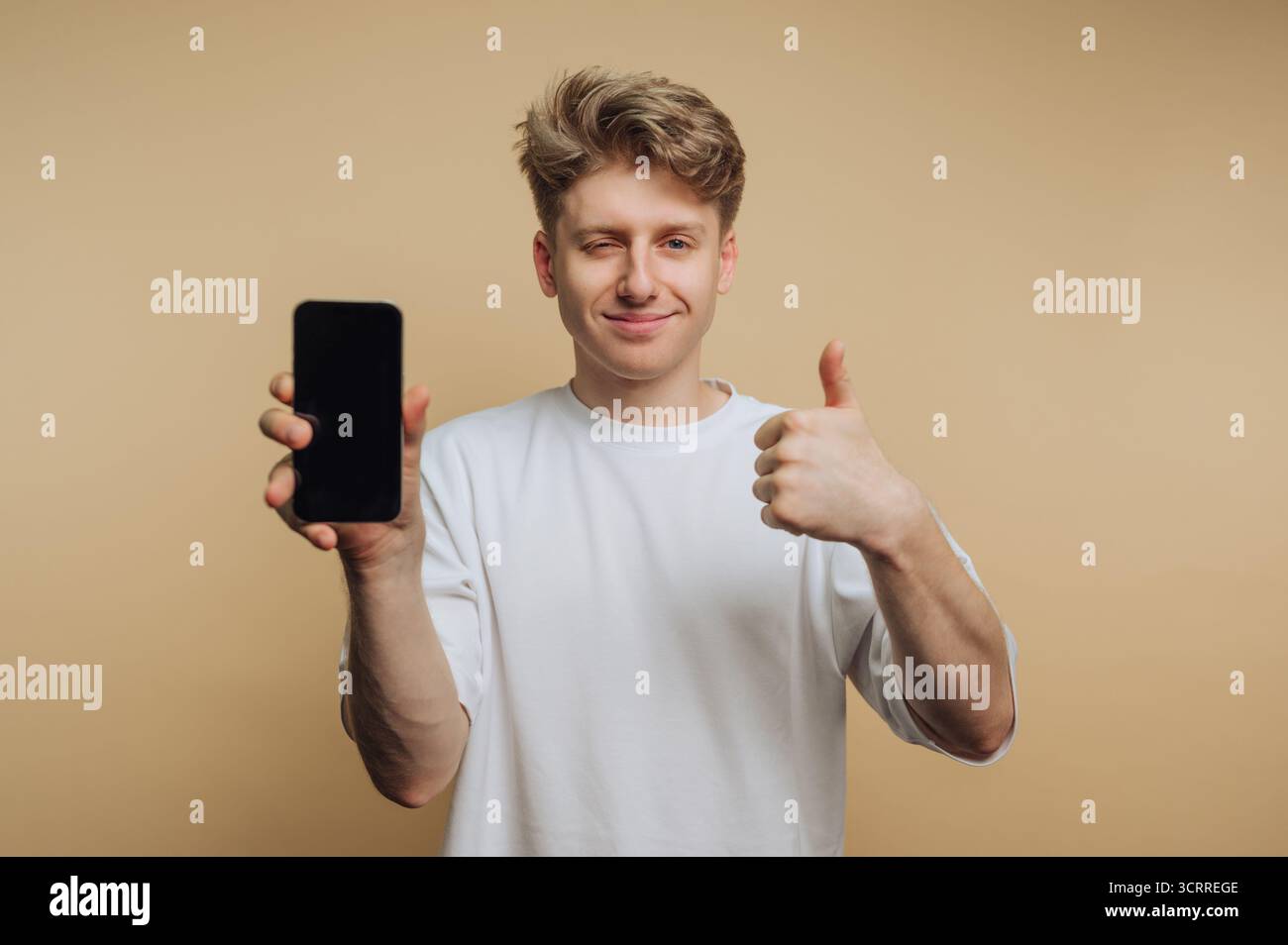 A young man with blonde hair confidently shows a smartphone while giving a thumbs up. He appears happy and engaged, against a warm neutral background. Stock Photo