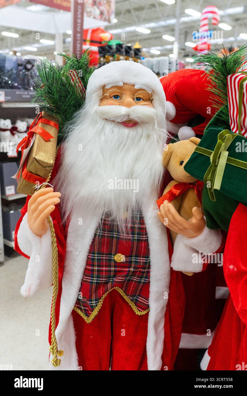 A decorative Santa Claus figurine stands in a store, holding a teddy bear and a gift, surrounded by other Christmas decor. Stock Photo