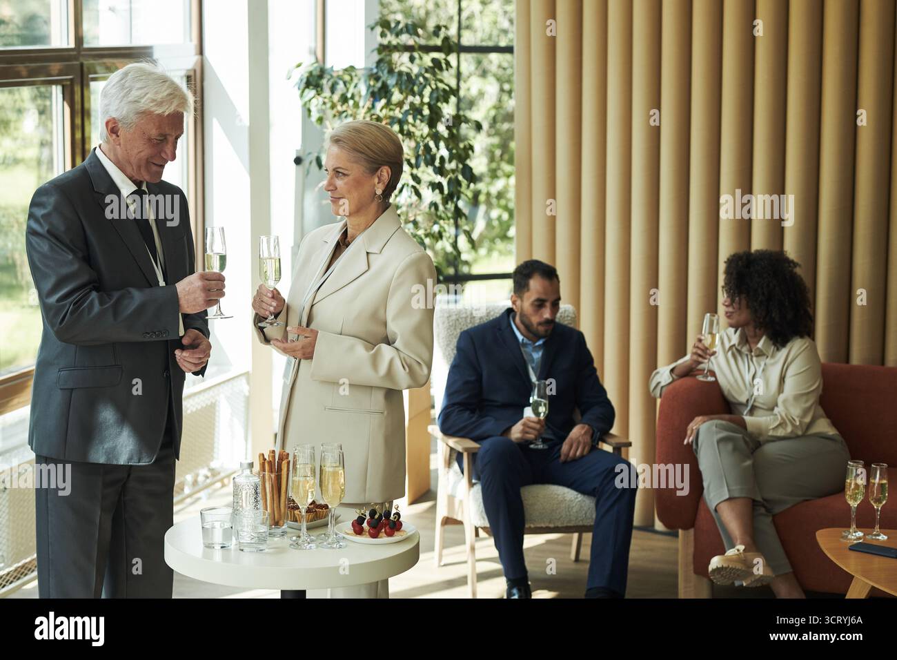 Senior Caucasian man and senior Caucasian woman standing and holding champagne glasses, talking near table with drinks, young Black woman and young Middle Eastern man sitting and drinking Stock Photo