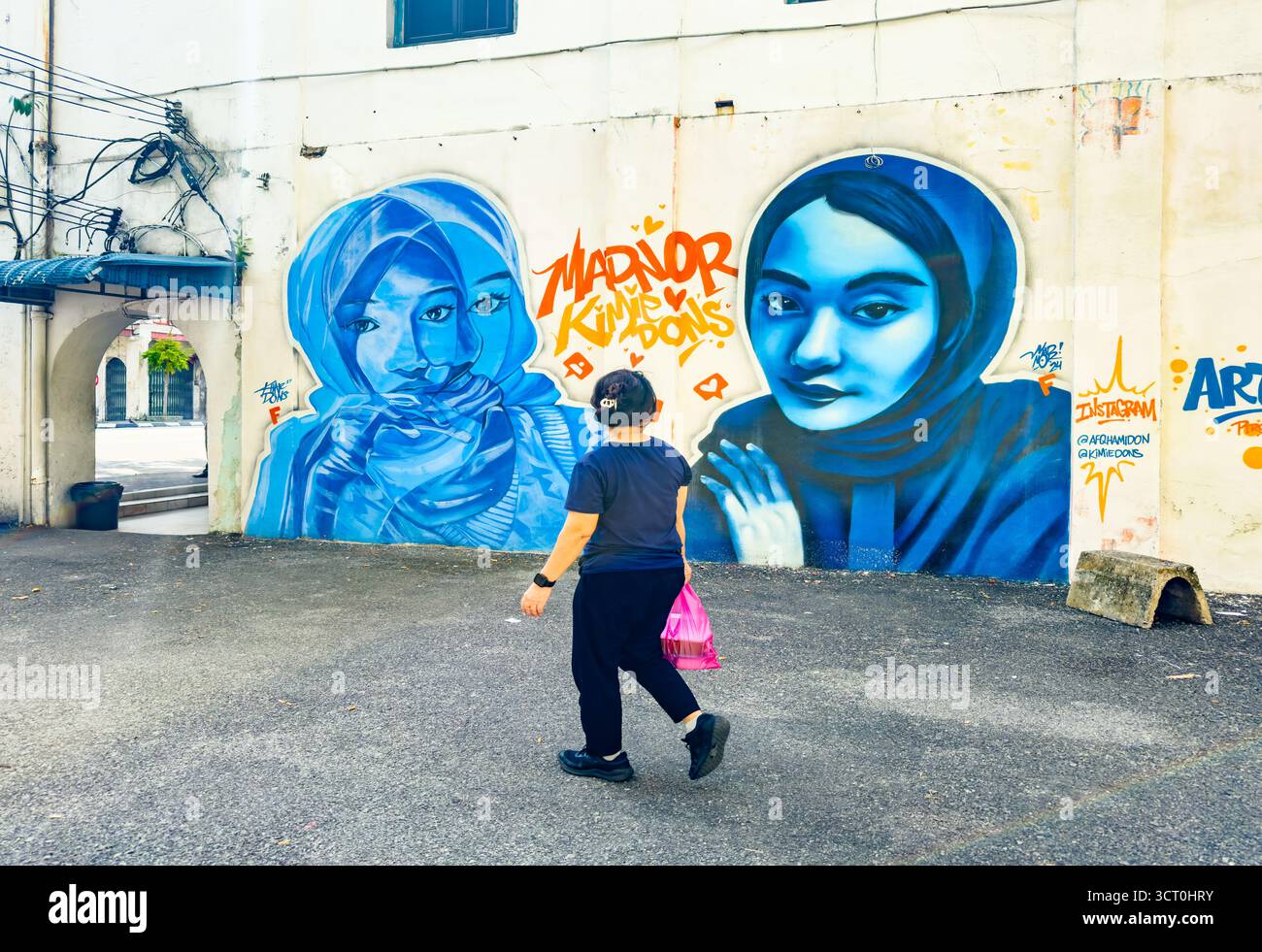 A person walks past a vibrant blue street art mural featuring two women in hijabs on a city wall ipoh malaysia - Smartphone Captured Stock Image