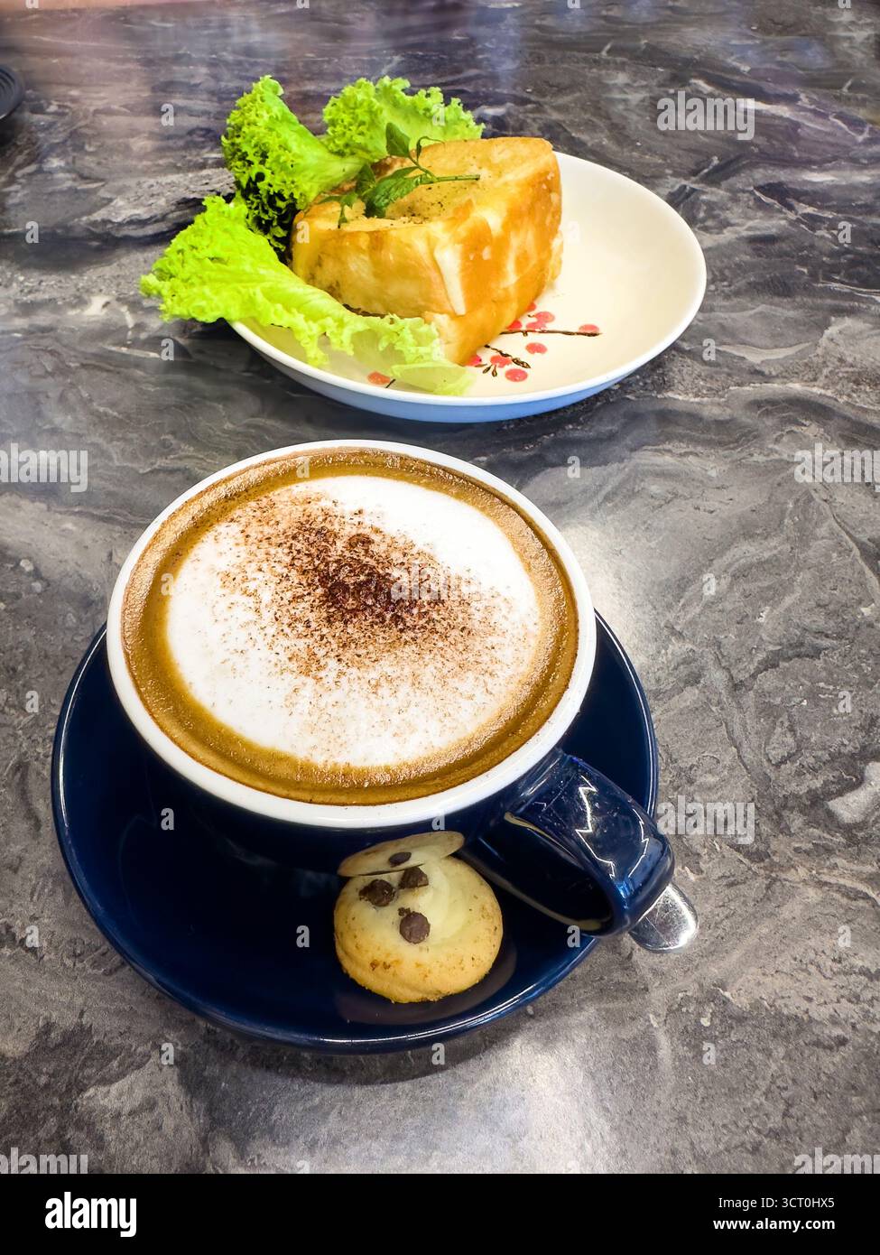 A frothy cappuccino with cinnamon served in a blue cup alongside a small biscuit on a marble table with a s kaya toast dish in background malaysia - Smartphone Captured Stock Image