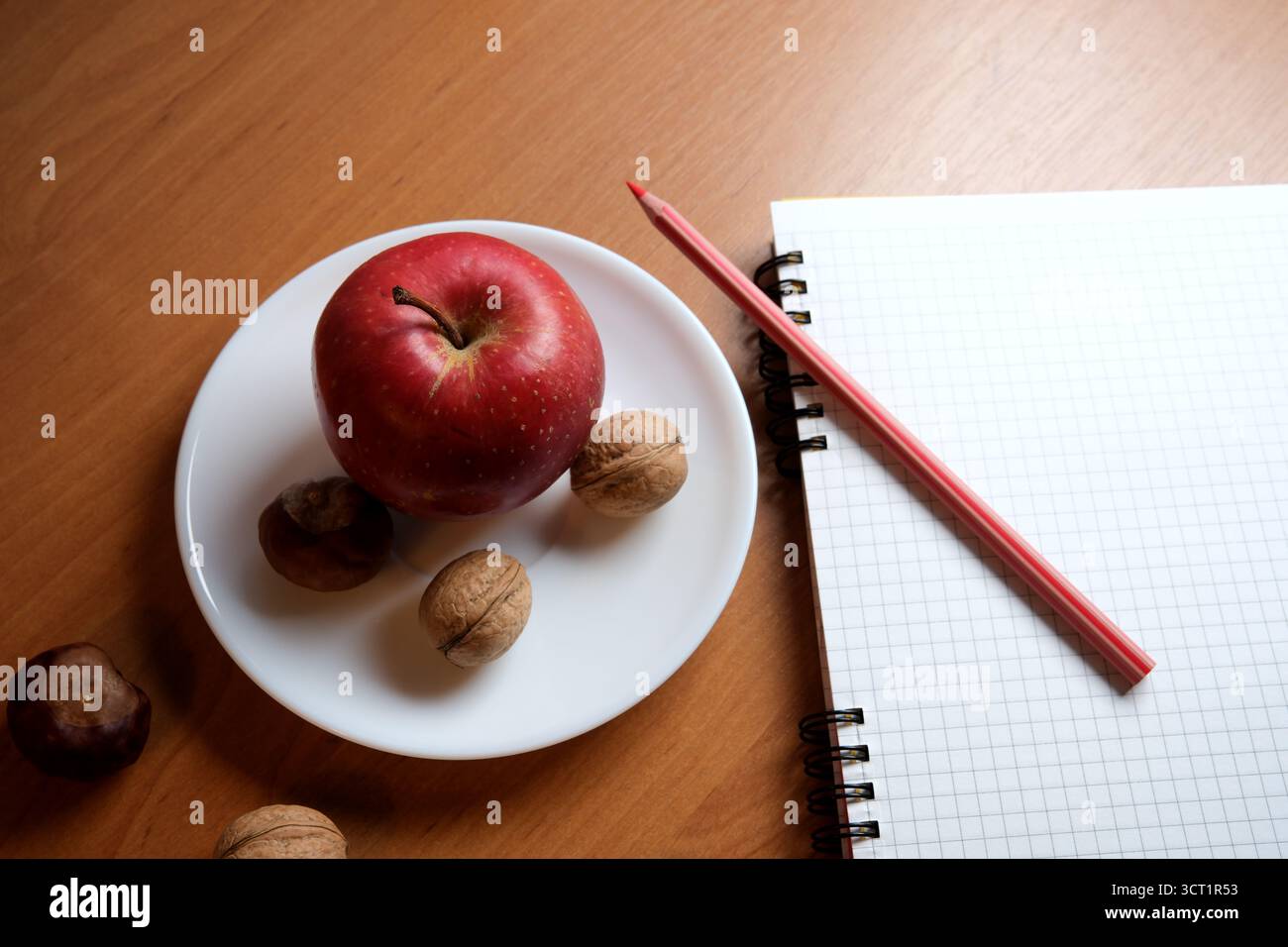 A bright red apple with walnuts sits on a plate near a notepad and pencil. Stock Photo