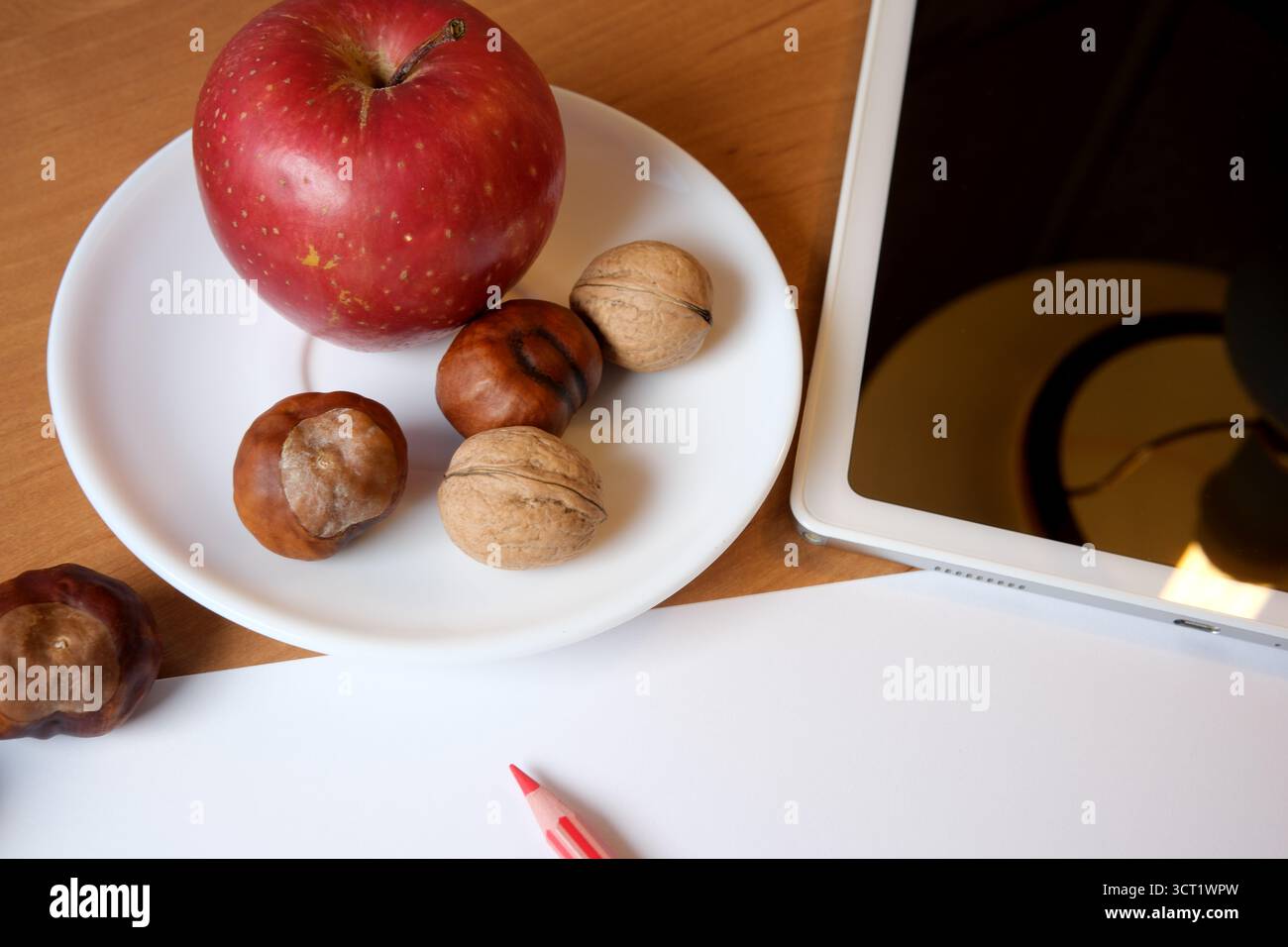 A red apple sits beside mixed nuts on a white plate next to a tablet and paper. Stock Photo