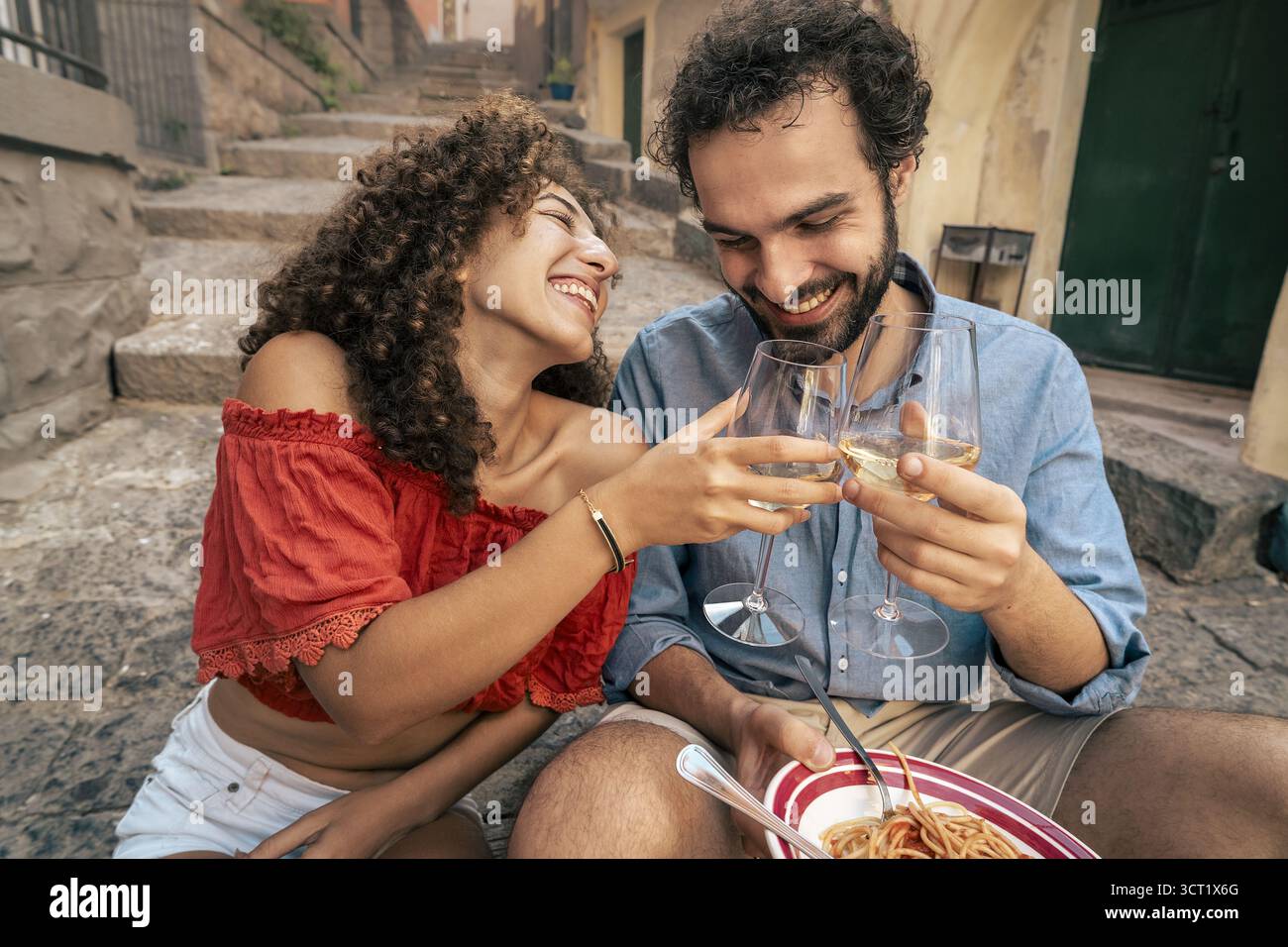 A happy young couple enjoys a romantic date, eating fresh pasta and toasting with white wine on the stone steps of a historic Italian alley. Stock Photo