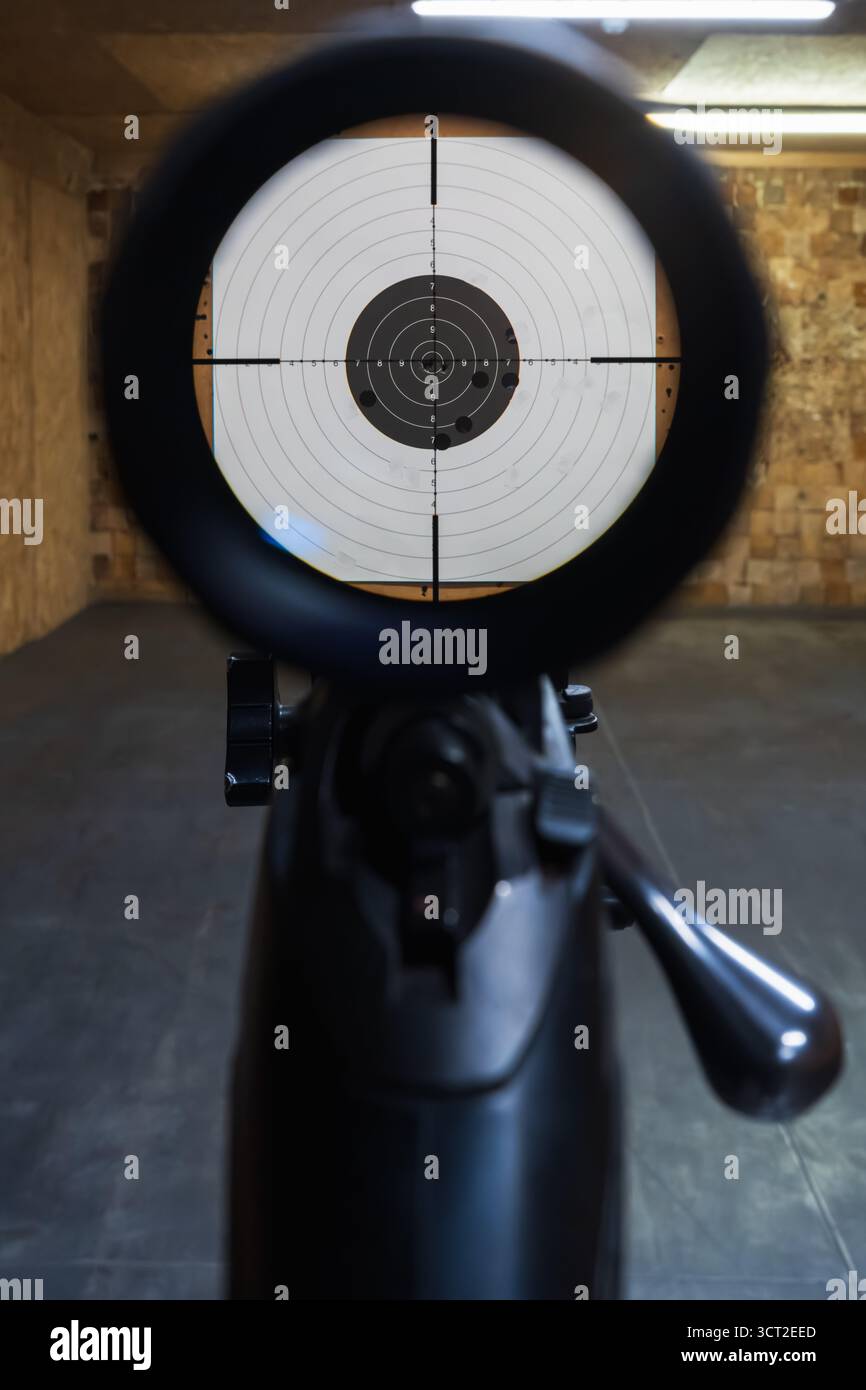 First-person view through a bolt-action rifle's telescopic sight, with crosshairs aimed at a paper target at an indoor range. Stock Photo