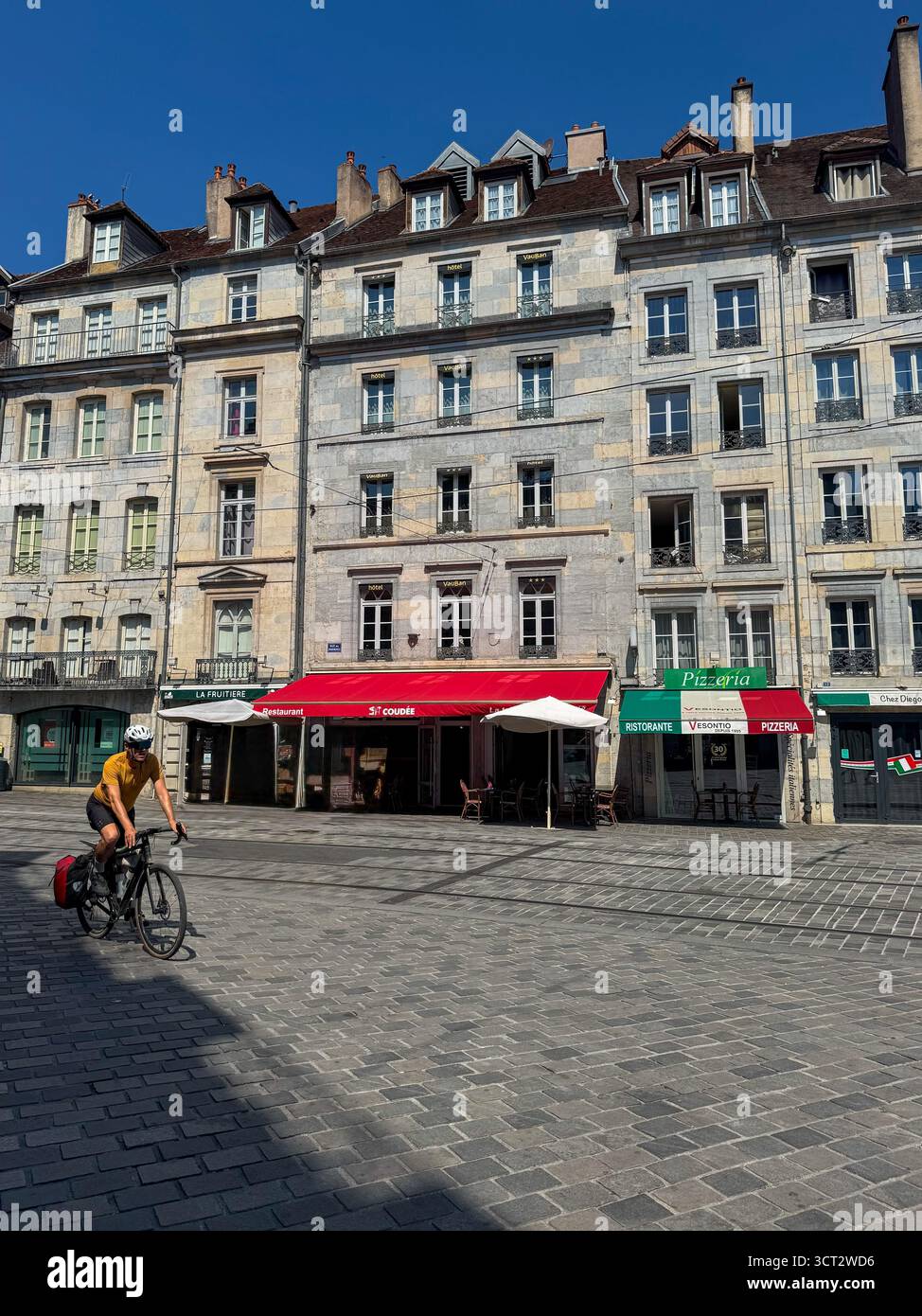 Cyclist in sportswear rides across a sunlit cobblestone square in Besancon, with historic stone building and red tiled roofs in the city center - Smartphone Captured Stock Image