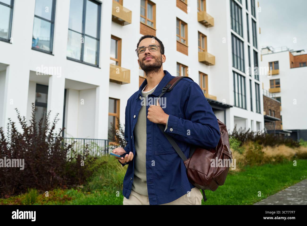Young Black Man Walking Outdoors Holding Smartphone and Backpack Stock Photo