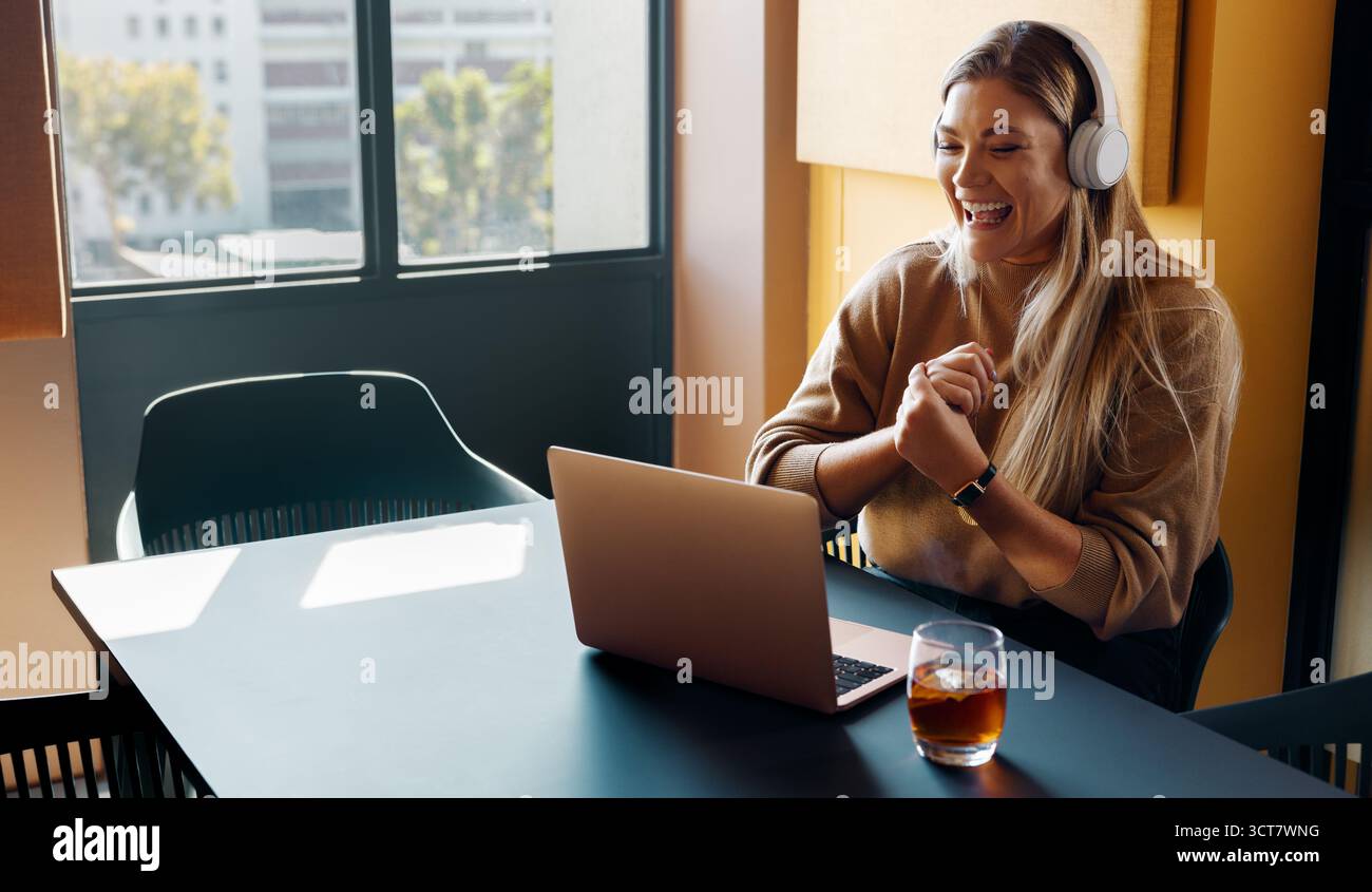 A smiling young woman wearing headphones having an engaging video call in a sunny workspace. The scene conveys professionalism, joy, and the modern ex Stock Photo