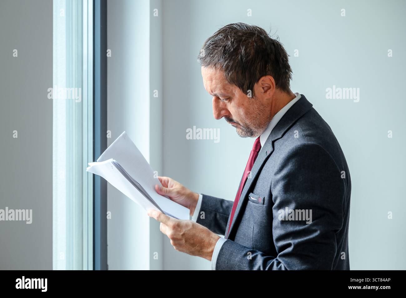 Businessman in formal suit reading documents near office window, focused professional corporate executive analyzing paper report in modern office envi Stock Photo