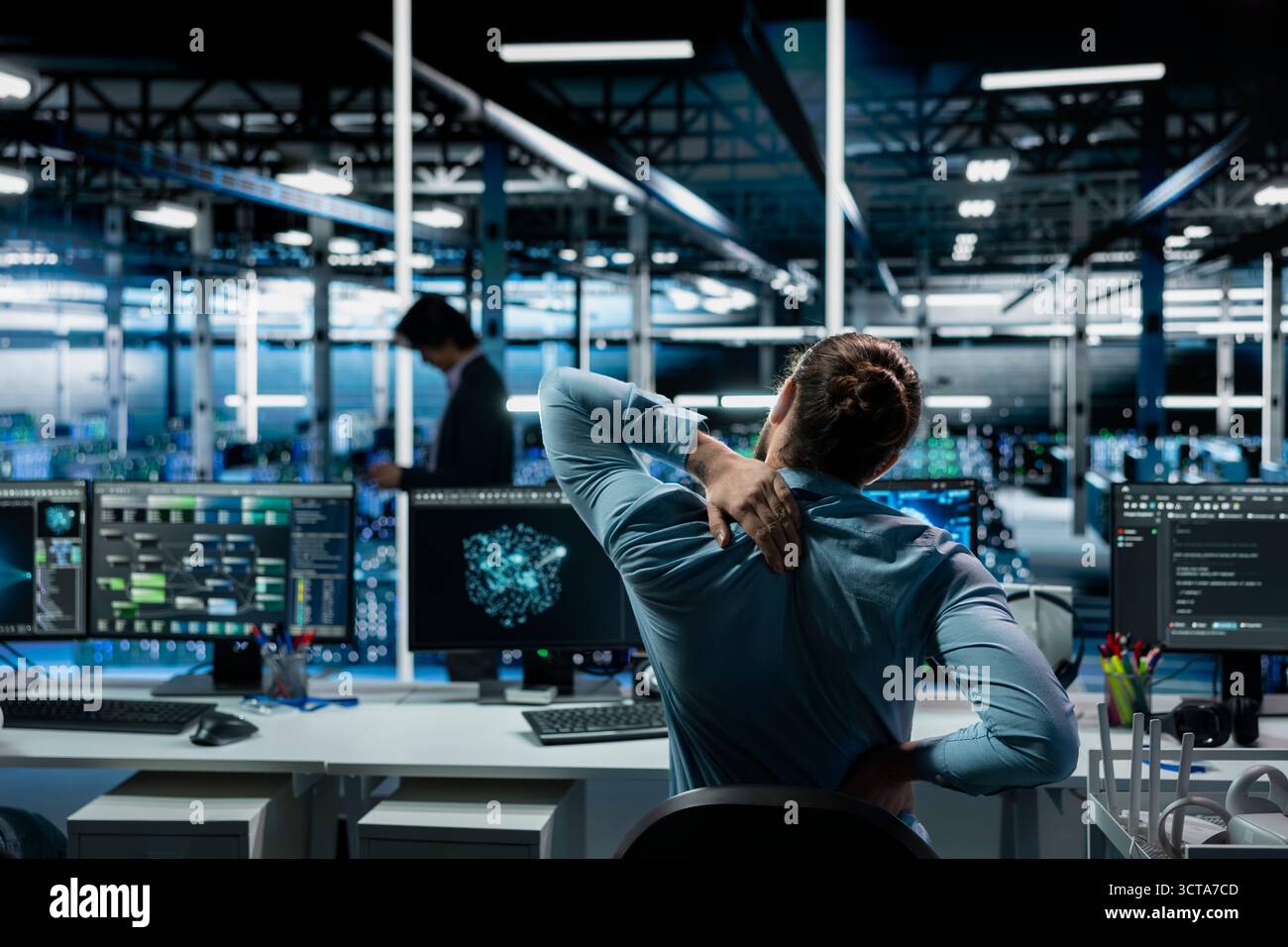 Tired data center software developer stretching to ease back tension while using AI LLM assistant. Server room IT professional massages injured back after long hours at computer coding with AI chatbot Stock Photo
