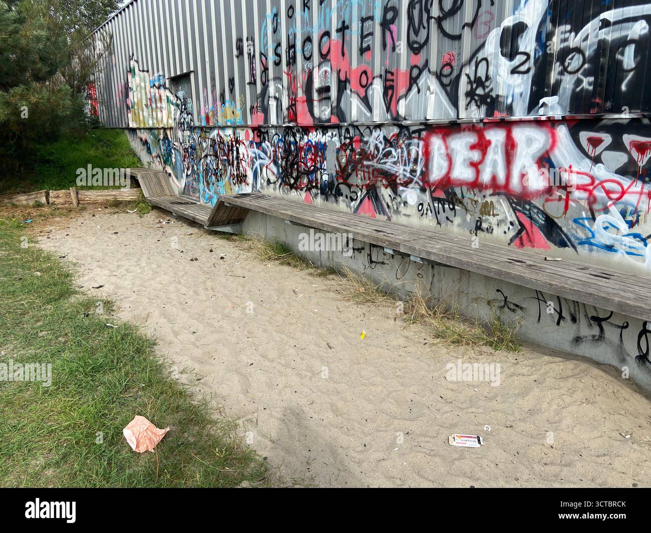 A colorful graffiti-covered wall with a bench attached, sand and patchy grass in front, scattered garbage on the ground adds urban grit. - Smartphone Captured Stock Image