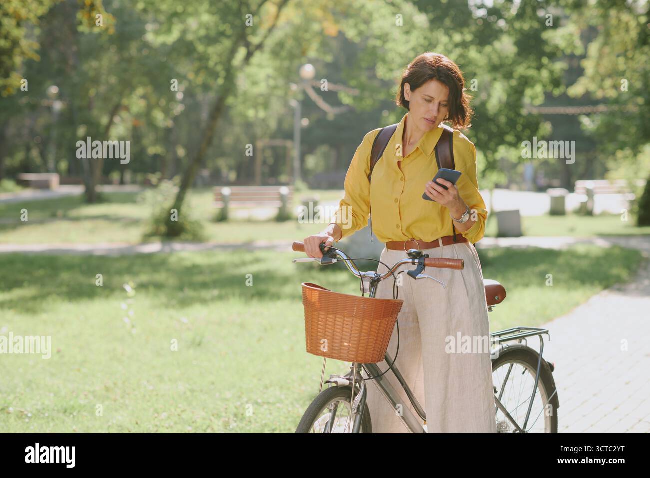 Caucasian Middle Aged Woman Walking Bicycle While Checking Smartphone Outdoors Stock Photo