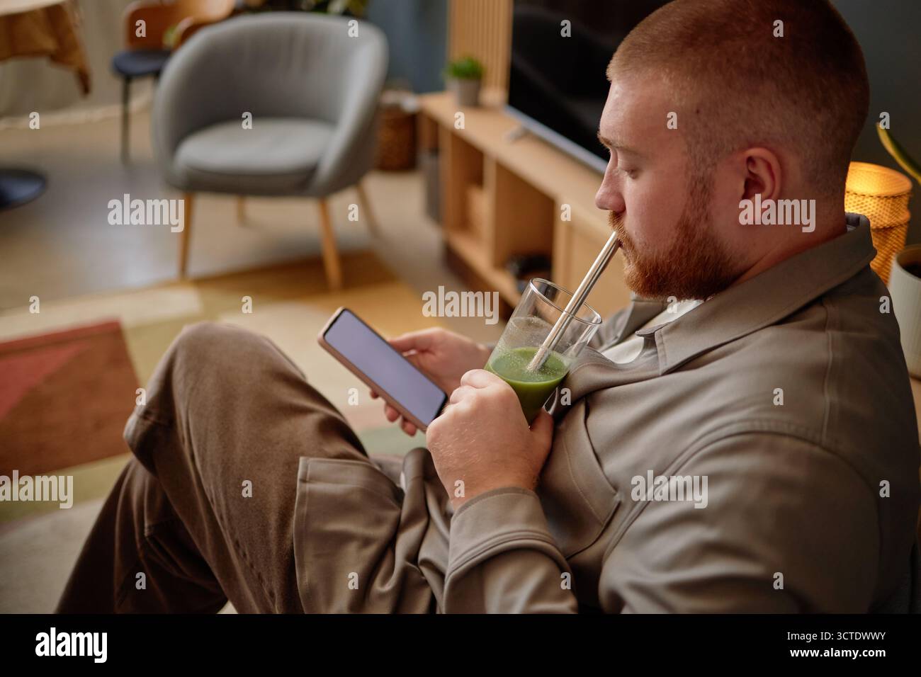 Young Adult Man Sitting Indoors Using Smartphone While Drinking Smoothie Stock Photo