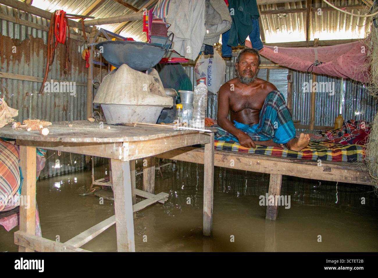 Rangpur, Bangladesh - 06 July 2024: View of a man sitting on a bed inside a corrugated metal home, surrounded by floodwaters reaching the table's surface. Stock Photo