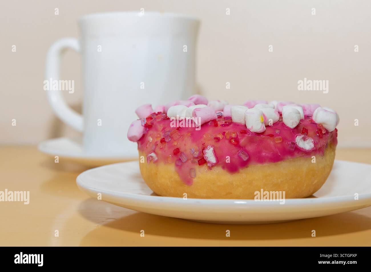 Donut with pink icing and marshmallows on plate with white cup on table. Sweet dessert for coffee break. Stock Photo