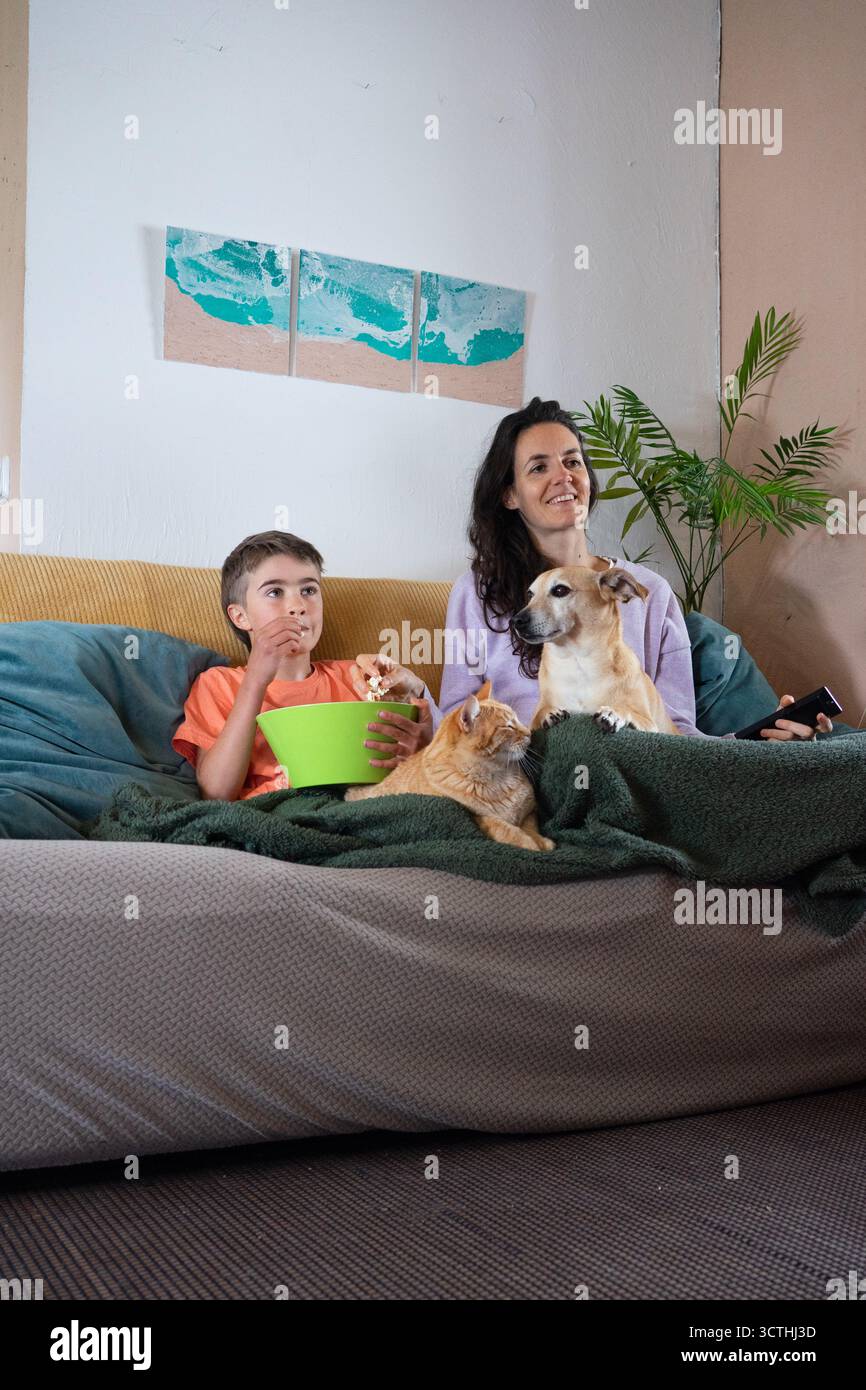 Happy family enjoying a movie night at home with their dog and cat, eating popcorn Stock Photo
