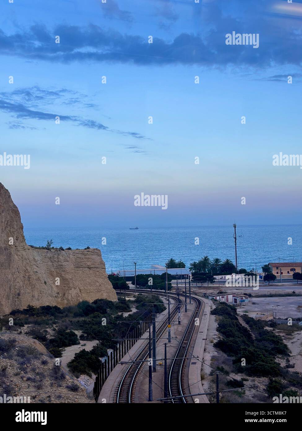 A scenic view overlooking a set of railway tracks that curve along the coast at dusk. To the left, a steep, sandy cliff descends toward the tracks. - Smartphone Captured Stock Image